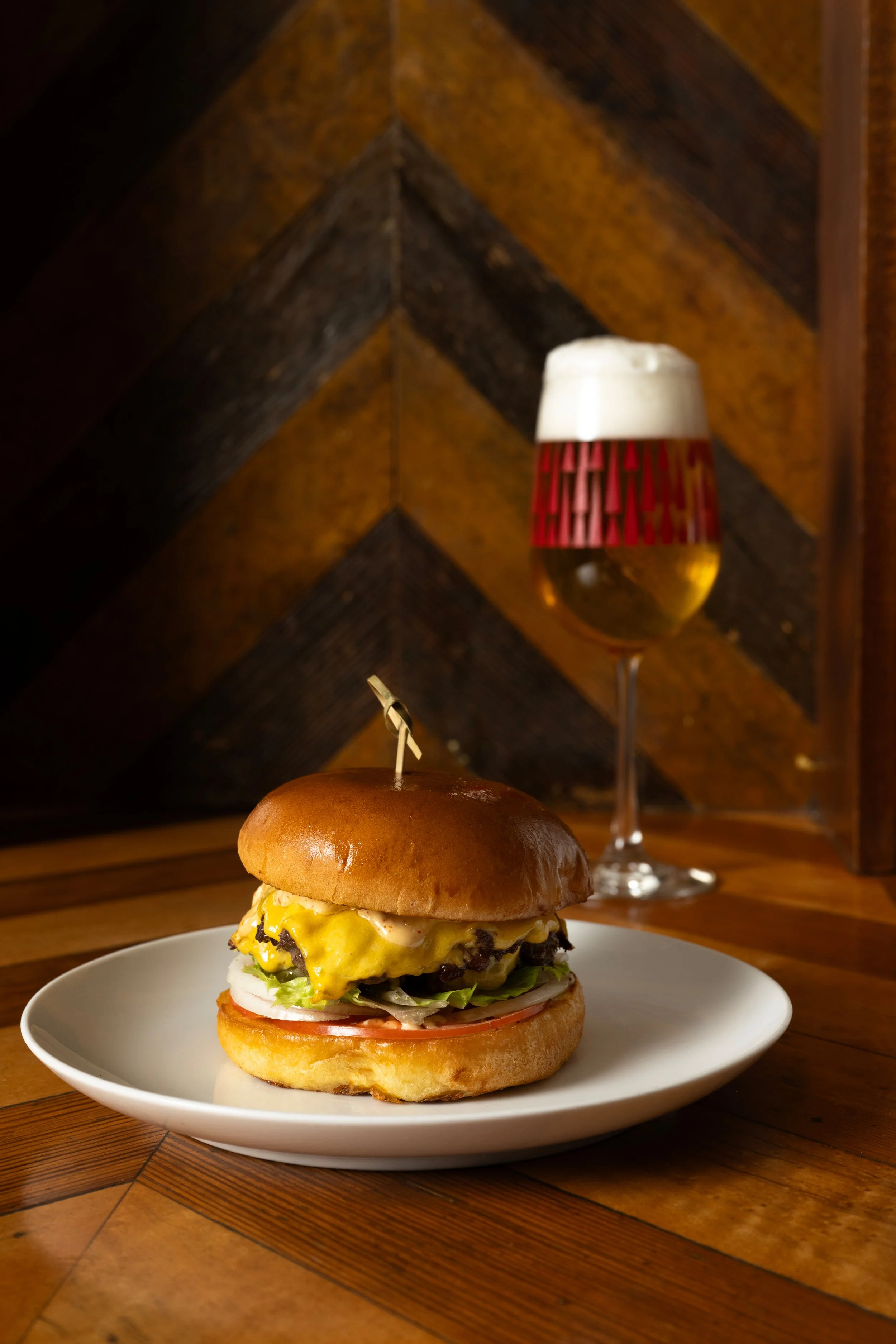 Smash burger with melted American cheese, lettuce, tomato, onion, and spicy mayo on a brioche bun alongside a patterned Tørst beer glass