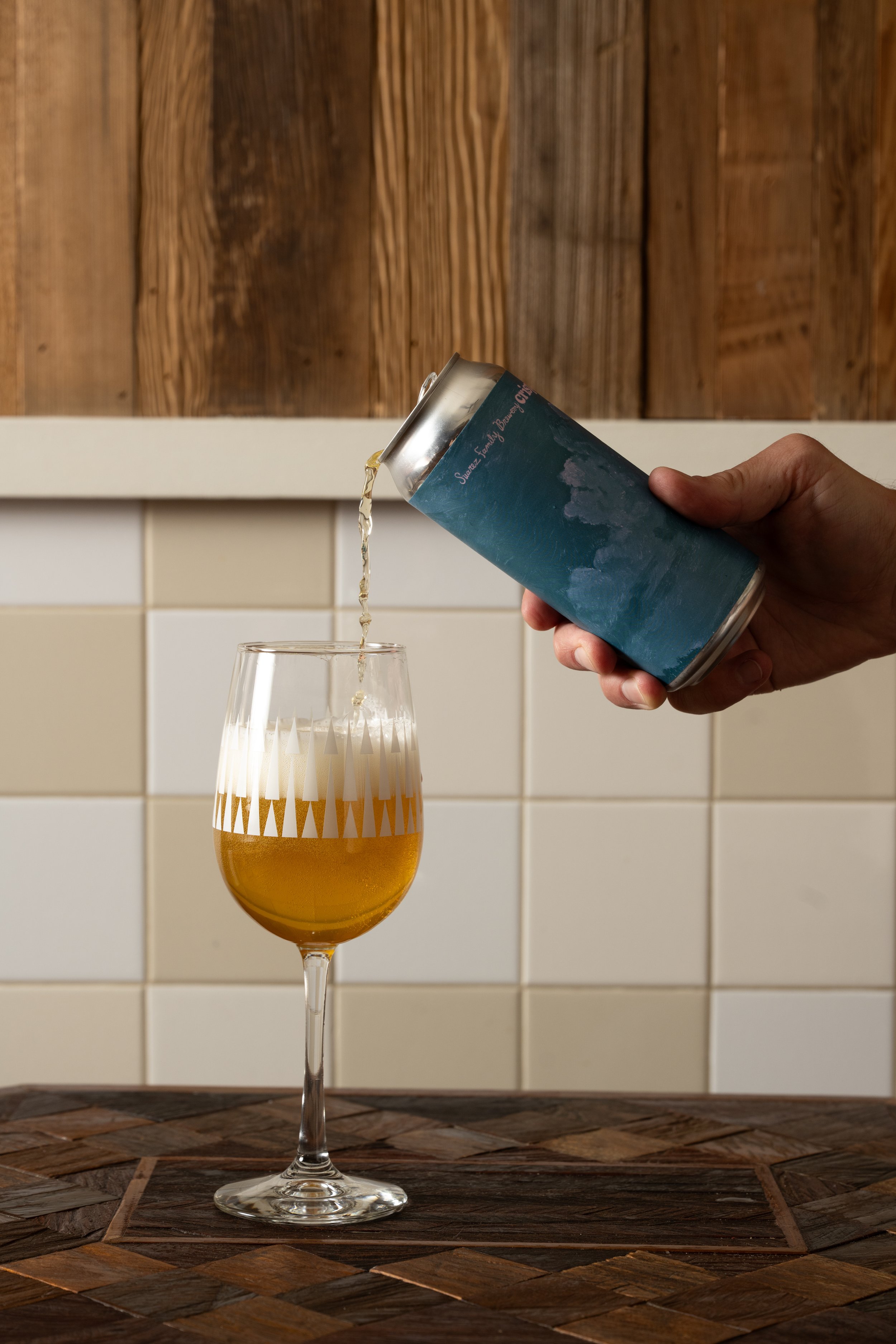 Can of beer being poured into a gold patterned Tørst glass on a wood-inlay table against white tile and wood panel walls