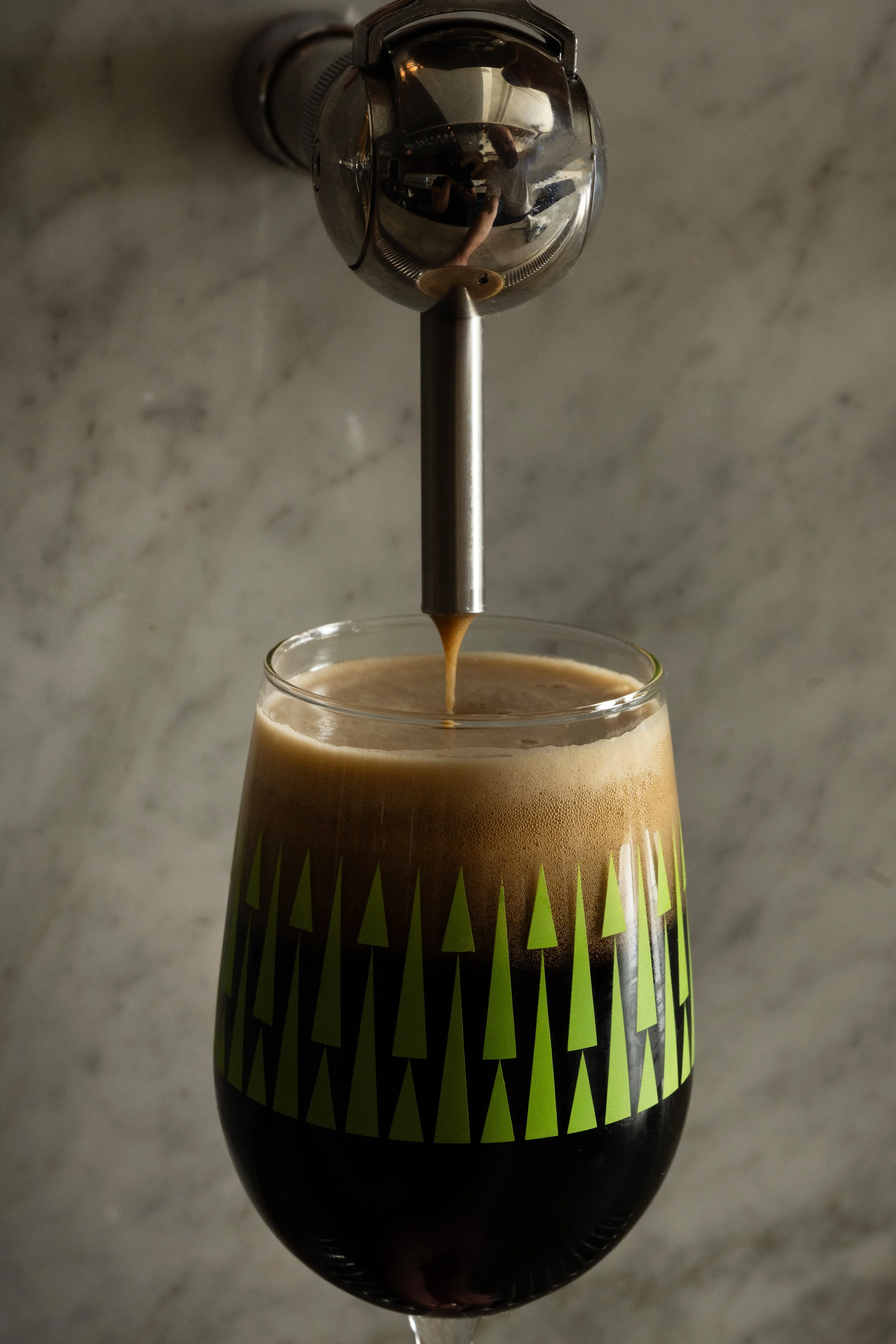 Close-up patterned Tørst beer glass filled with a dark stout and creamy foam head, sitting on the marble bar in front of draft tap handles