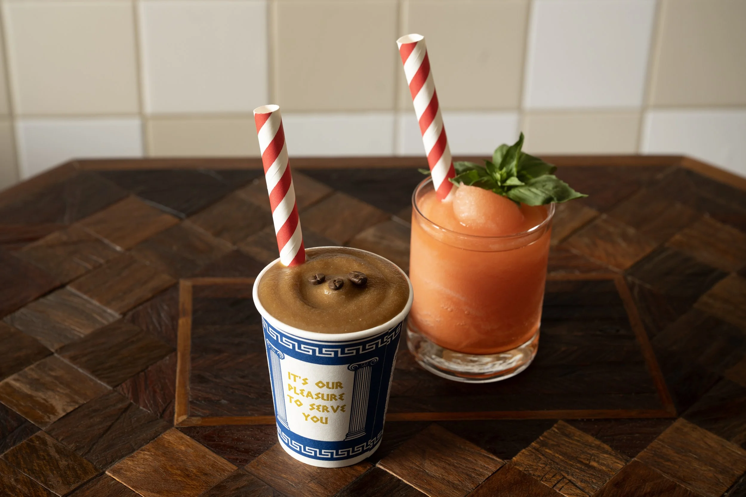 Frozen espresso martini in a paper cup and a frozen strawberry rhubarb cocktail with basil garnish, served with striped paper straws on a wood-inlay table
