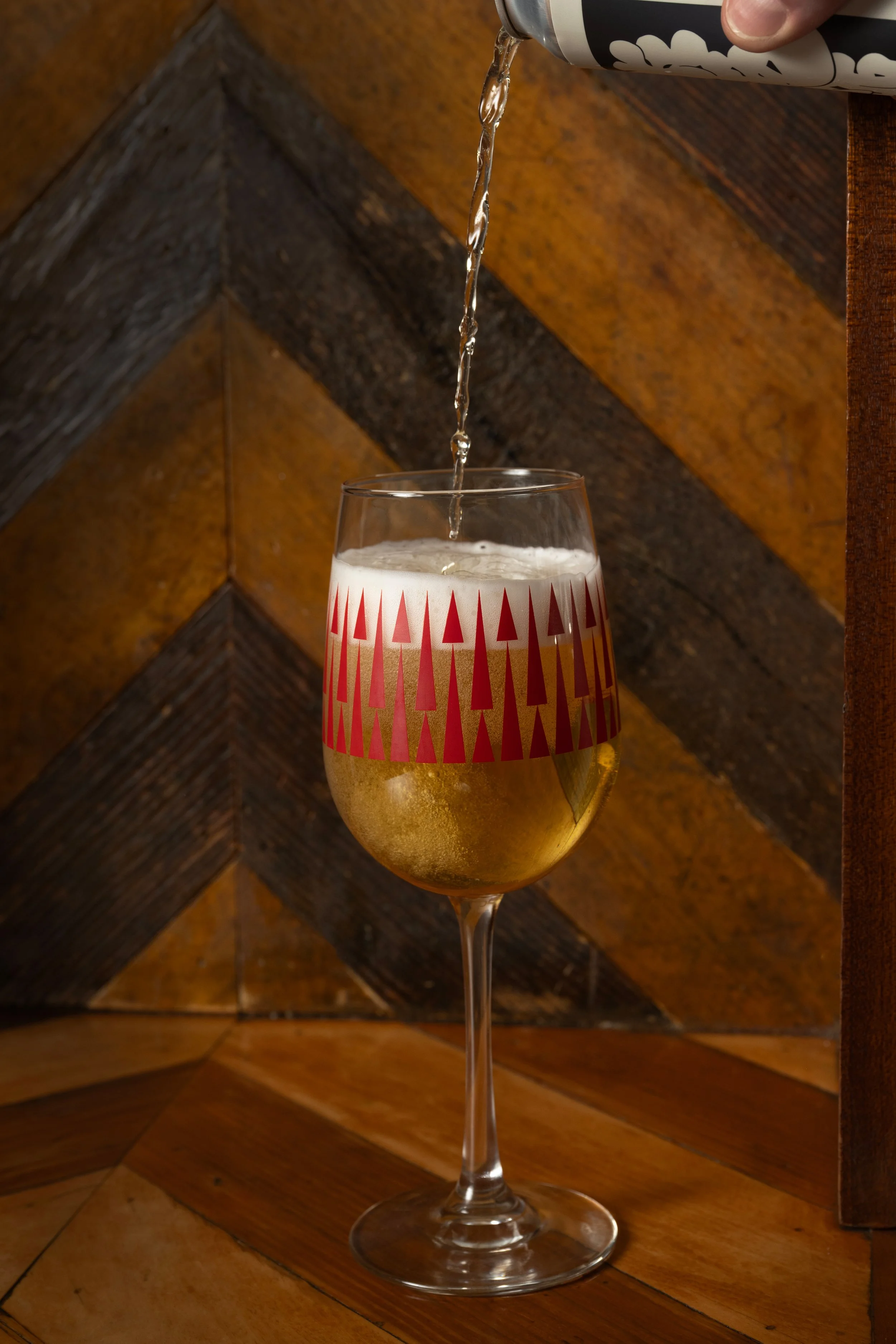 Beer being poured from a can into a red patterned Tørst glass against a herringbone wood wall