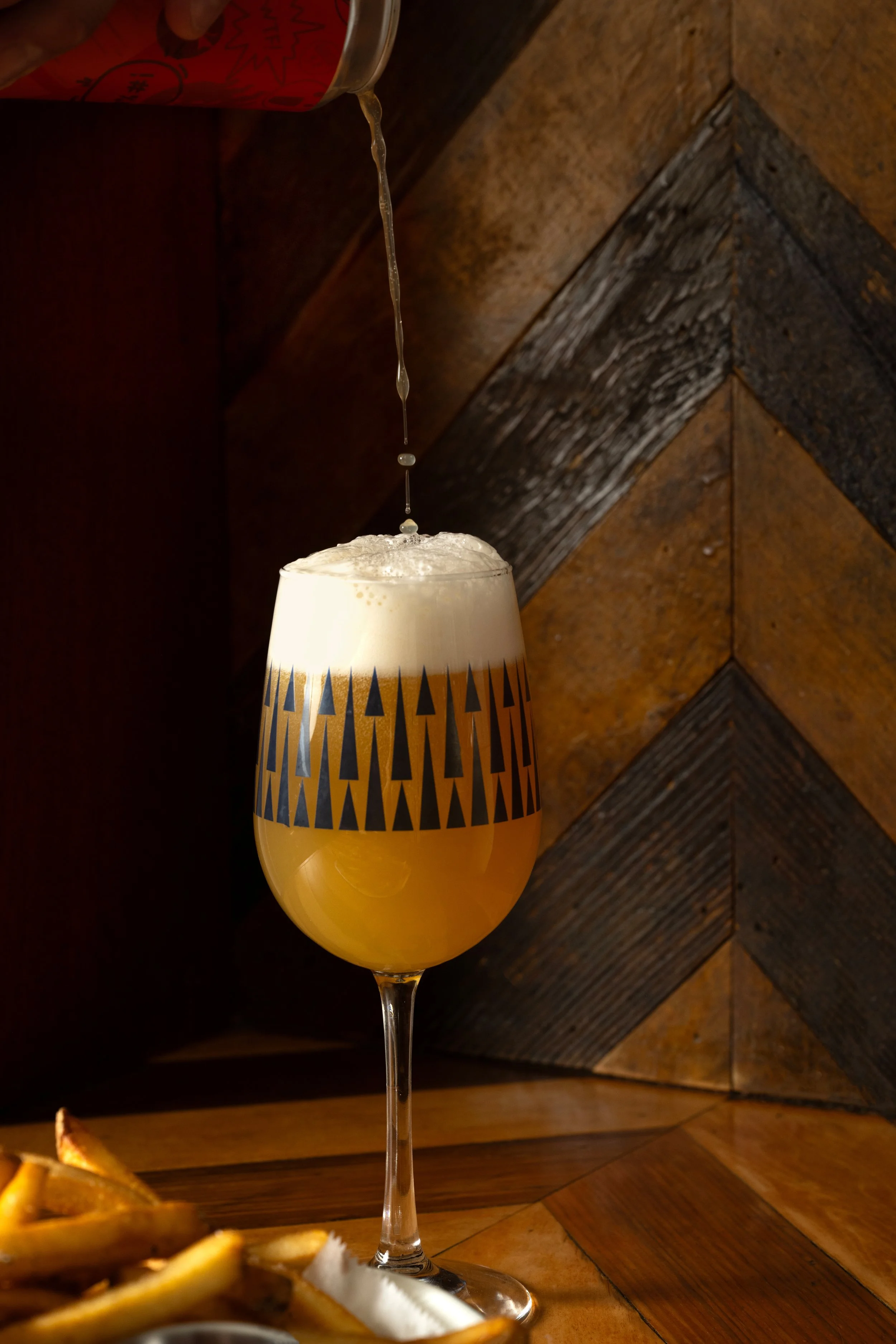Beer being poured into a patterned Tørst glass against a herringbone wood backdrop, with fries in the foreground