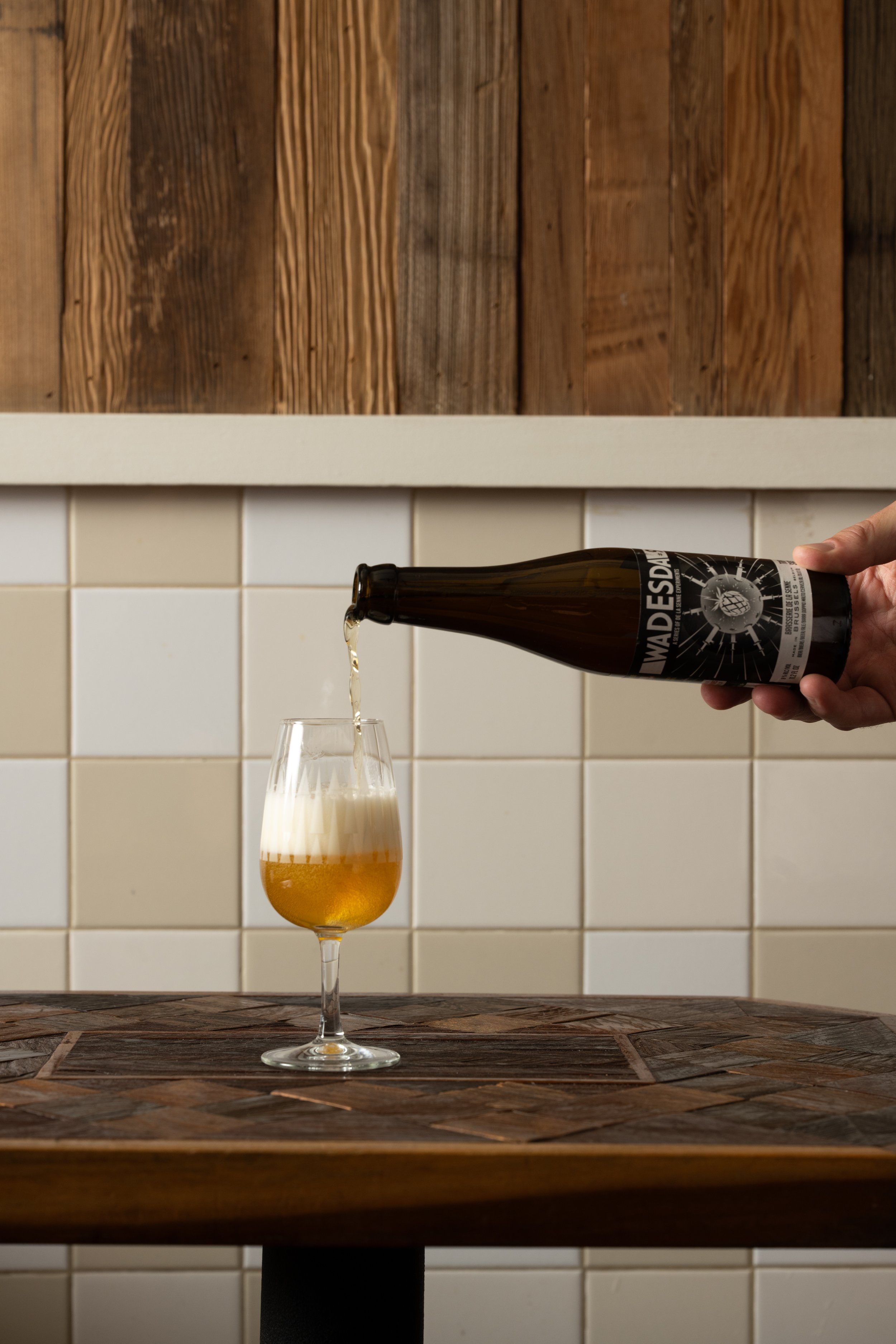 Bottle of beer being poured into a glass on a wood-inlay table against white tile and wood panel walls