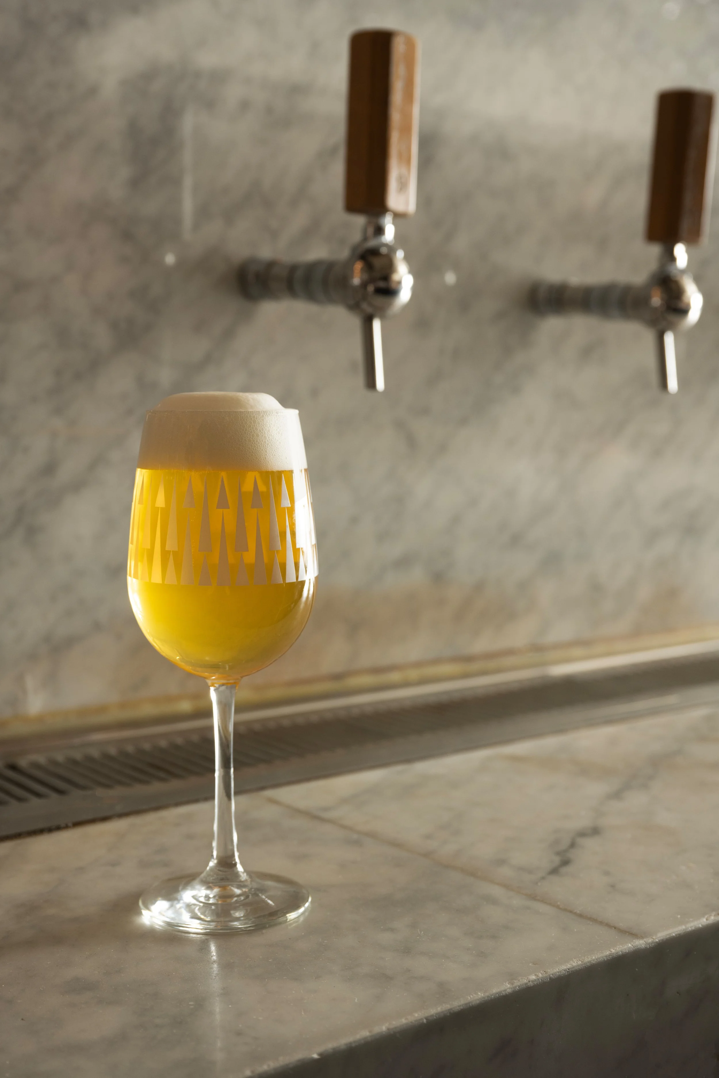 Patterned Tørst beer glass with a perfect head of foam resting on the marble bar with two wooden draft tap handles in the background