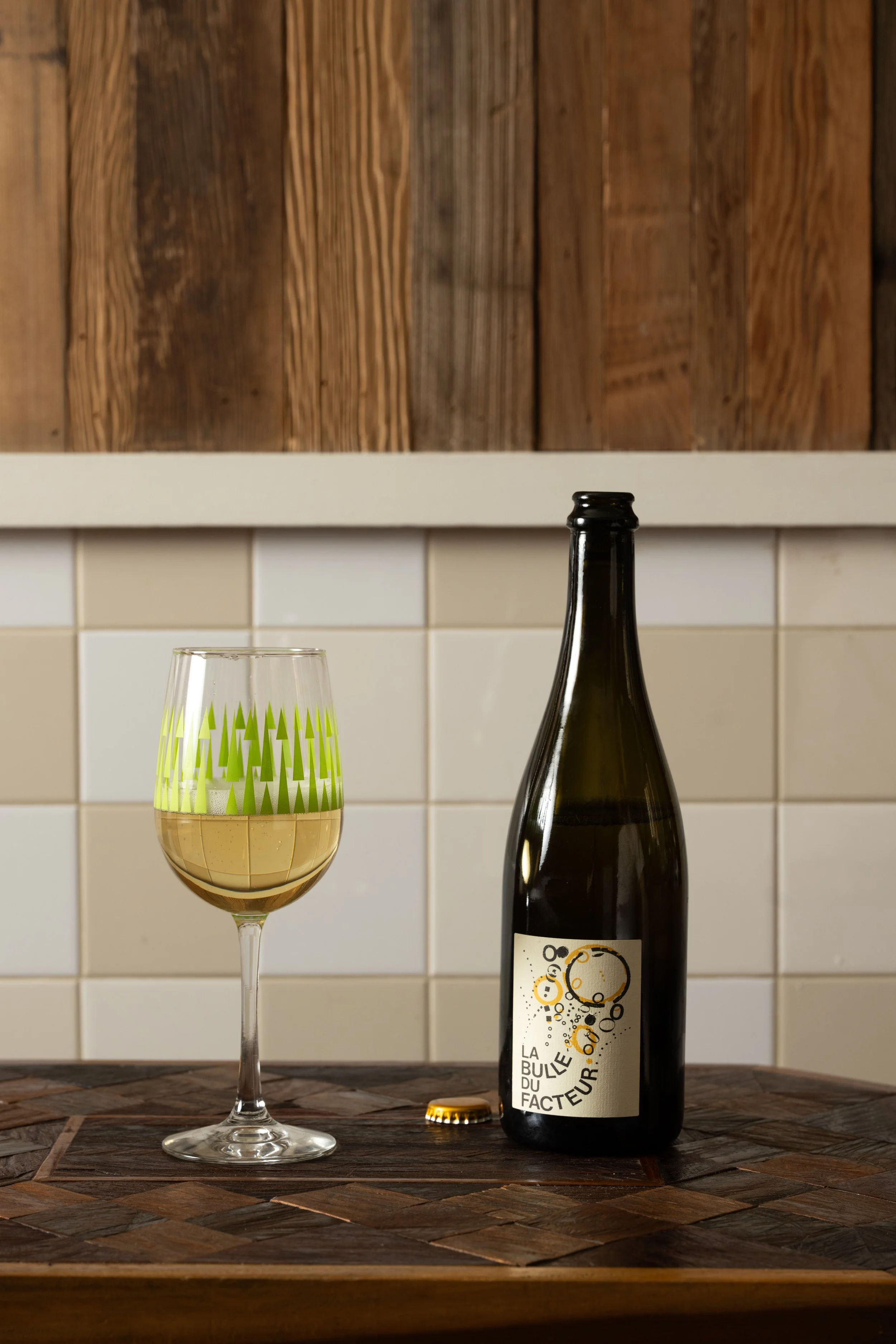 Bottle of beer alongside a poured glass of golden beer in a green patterned Tørst glass on a wood-inlay table