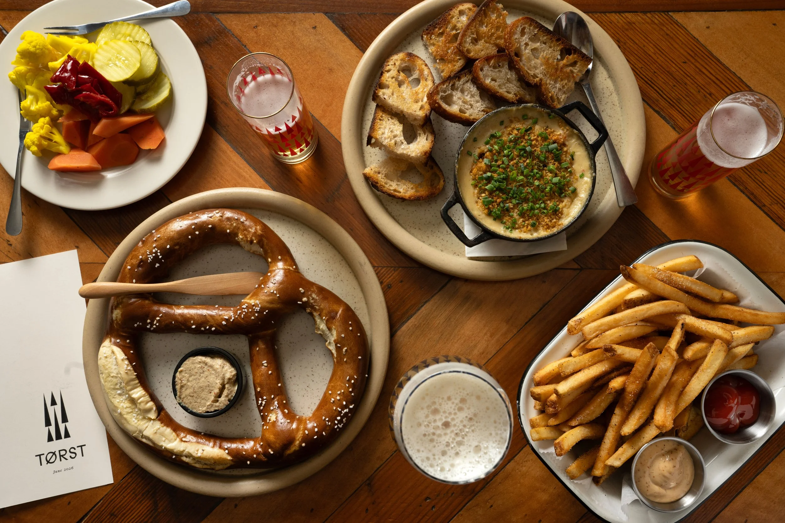 Overhead spread of Tørst bar food including a giant Bavarian pretzel, hot crab dip with sourdough, Kennebec fries, and pickled vegetables, alongside patterned beer glasses and the Tørst food menu