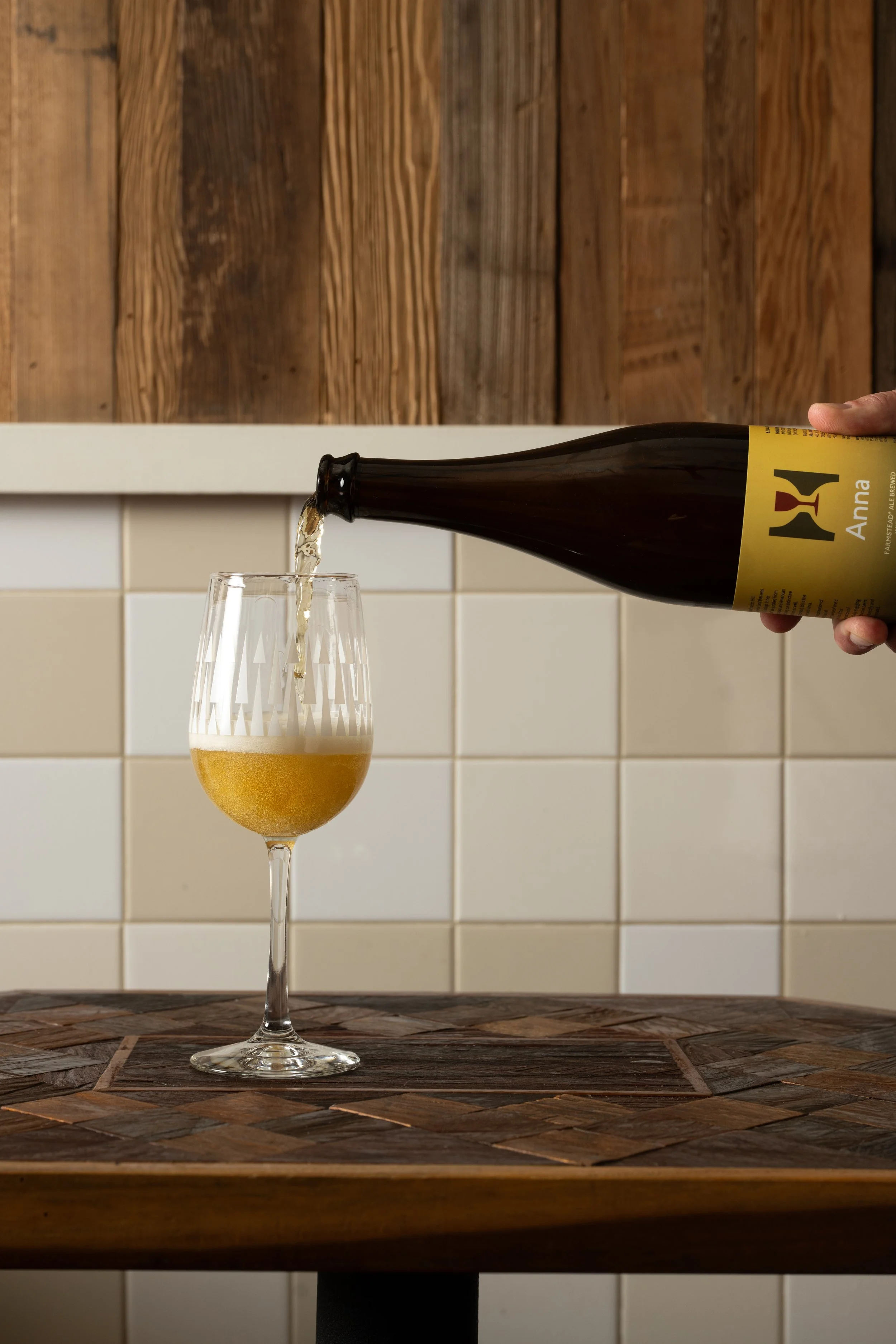 Craft beer being poured from a bottle into a clear patterned Tørst glass on a wood-inlay table against white tile and wood panel walls