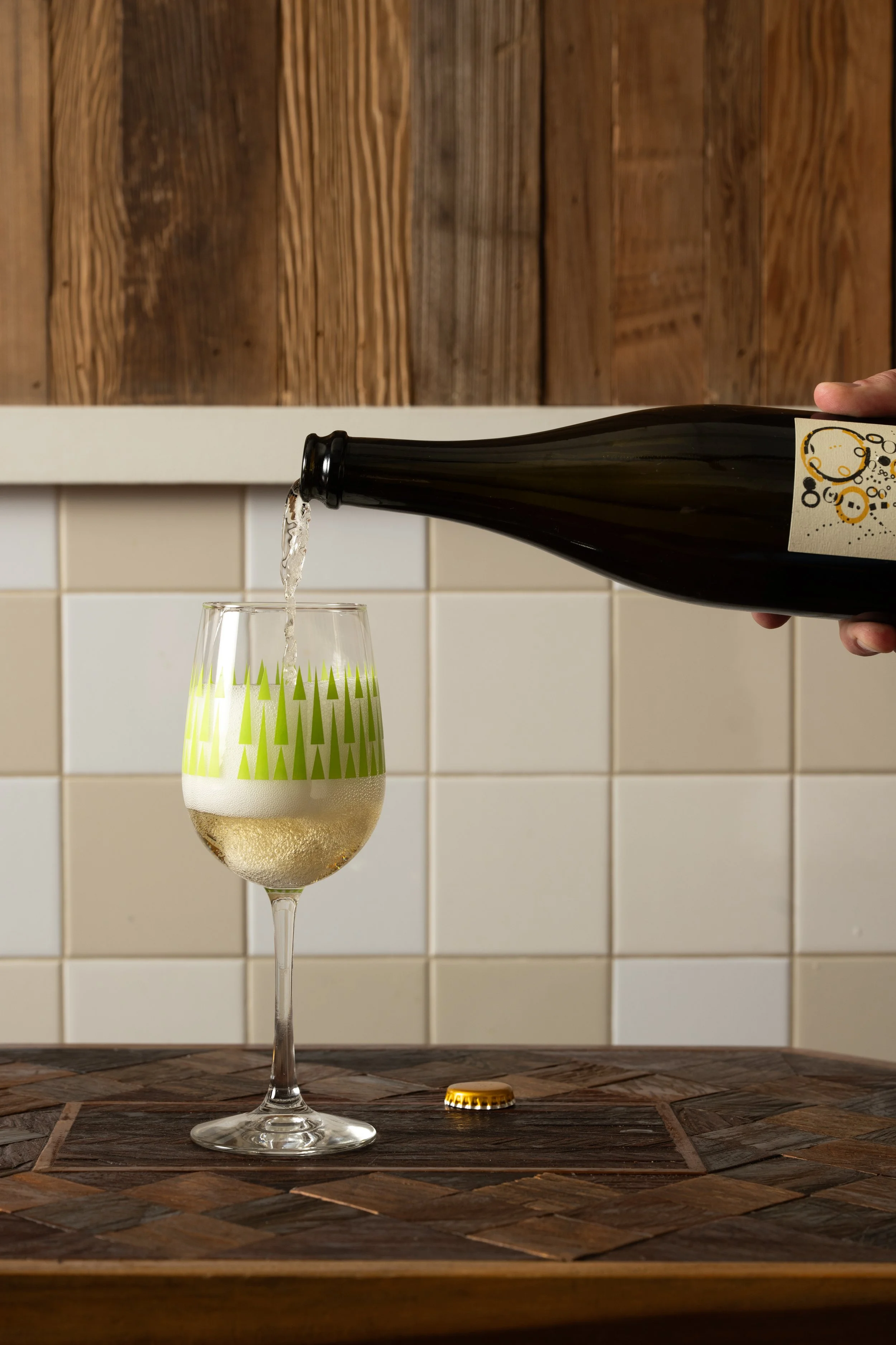 Craft beer being poured from a bottle into a green patterned Tørst glass on a wood-inlay table against white tile and wood panel walls