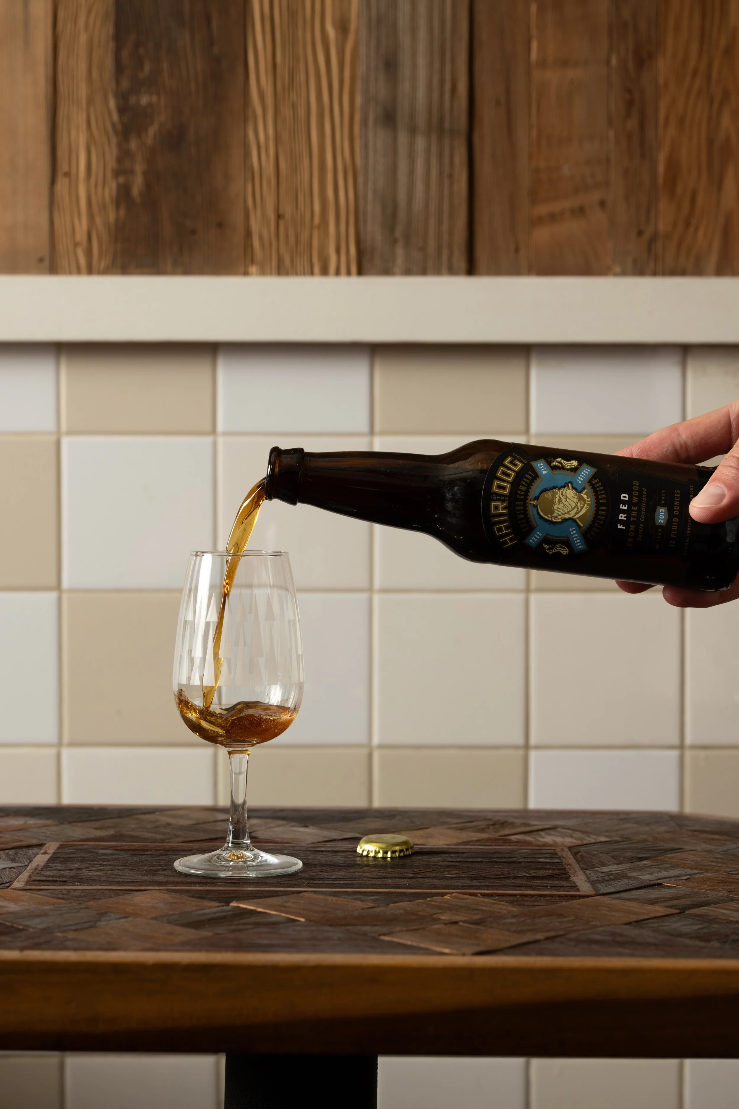 Beer being poured from a bottle into a glass on a wood-inlay table, bottle cap resting beside it
