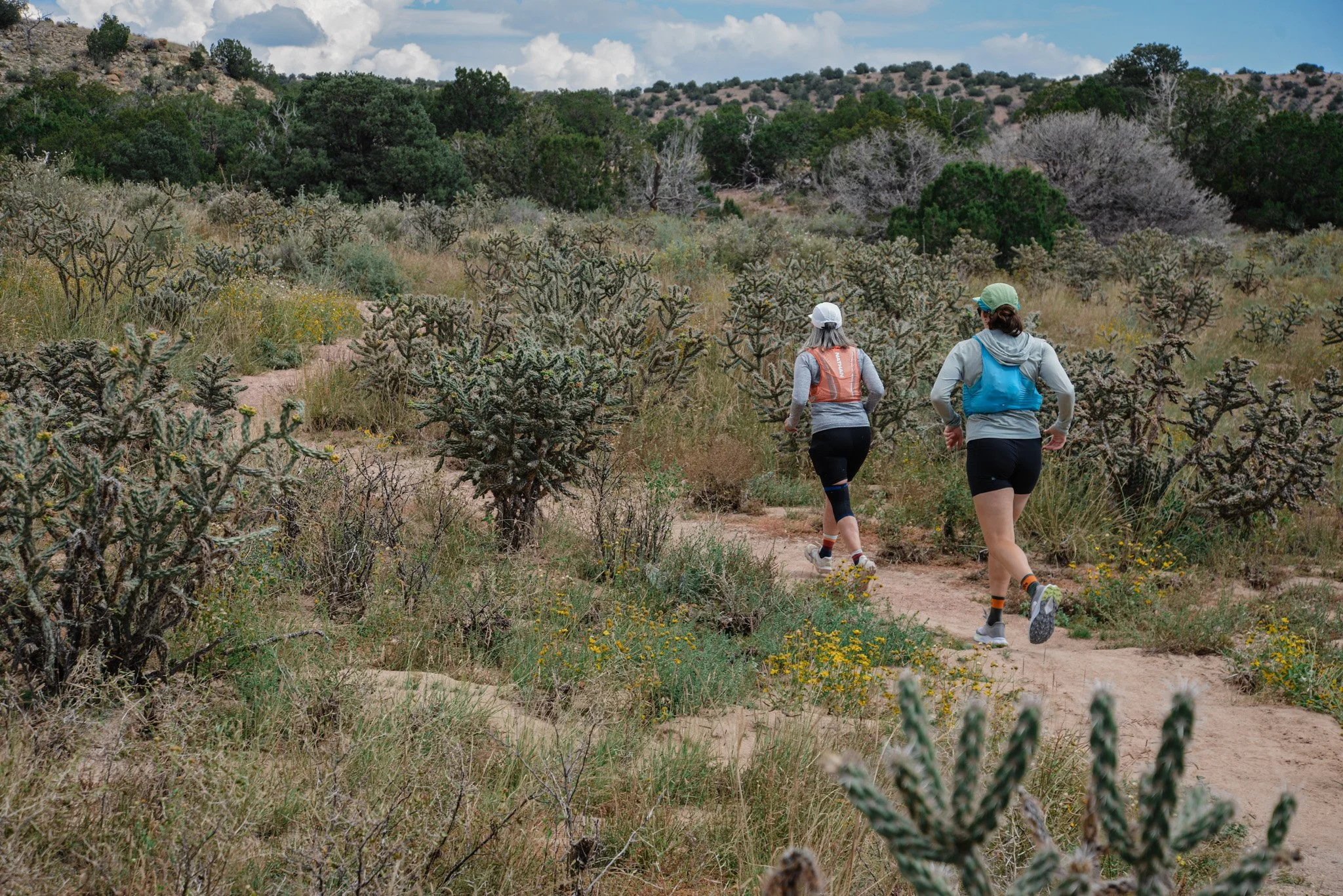 Two women trail running through a desert landscape with cacti and sparse vegetation under a partly cloudy sky.