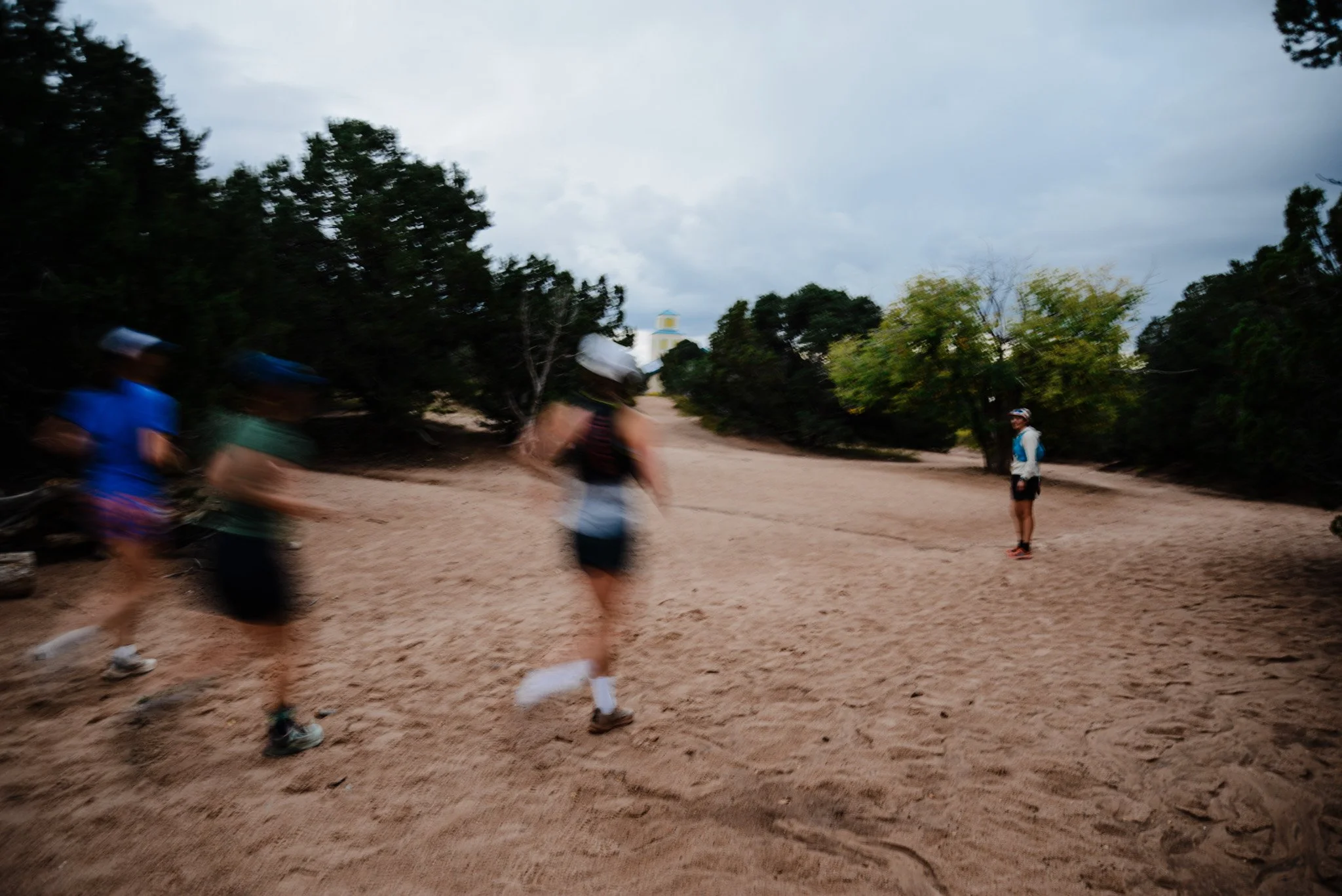 Blurred image of people walking on a dirt trail surrounded by trees, with an overcast sky and a small structure with a yellow roof in the distance.