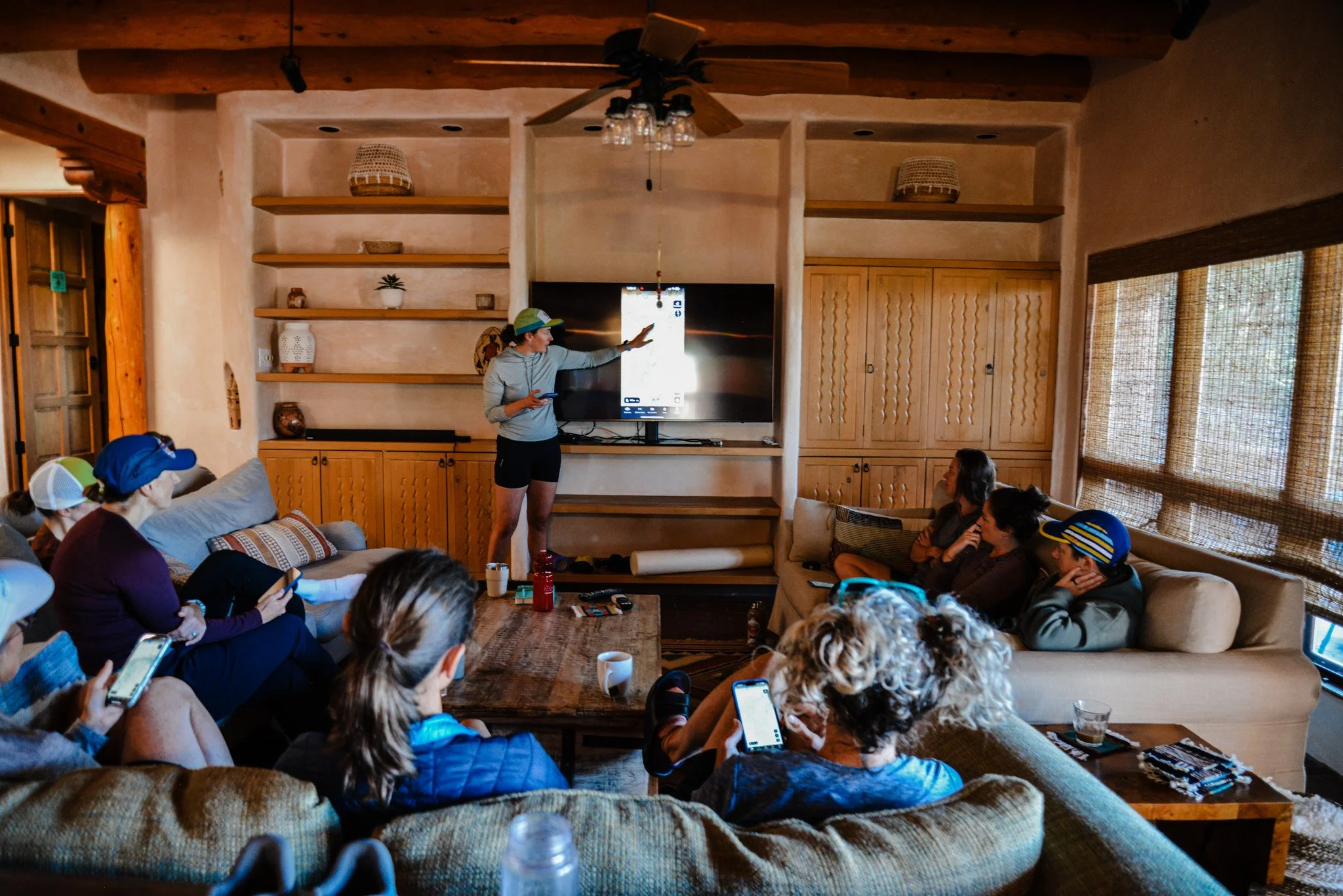 People seated on couches and chairs in a cozy living room, watching a woman stand in front of a large flat-screen TV showing a presentation, while some audience members hold phones.