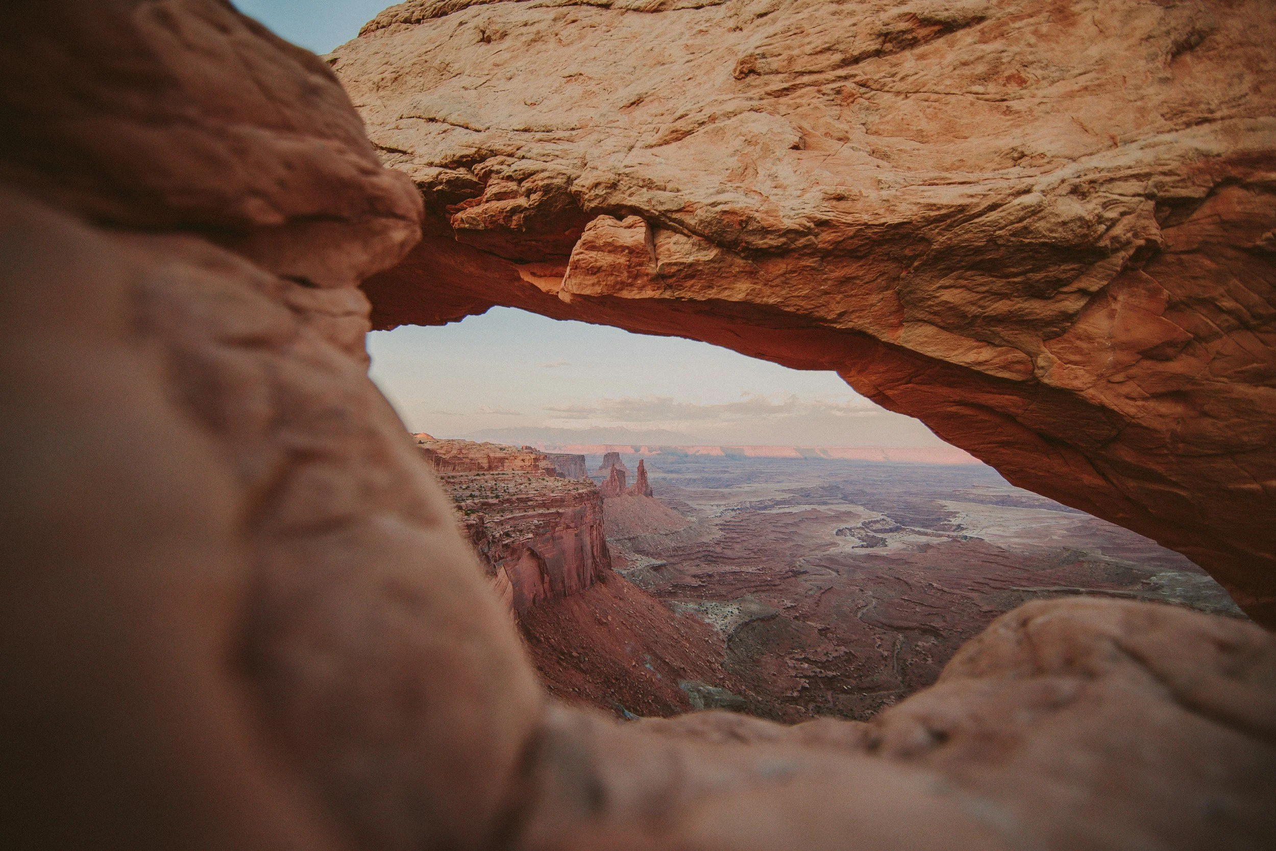 A view through a natural rock arch at the Grand Canyon, showing layered red rock formations and a distant river below.