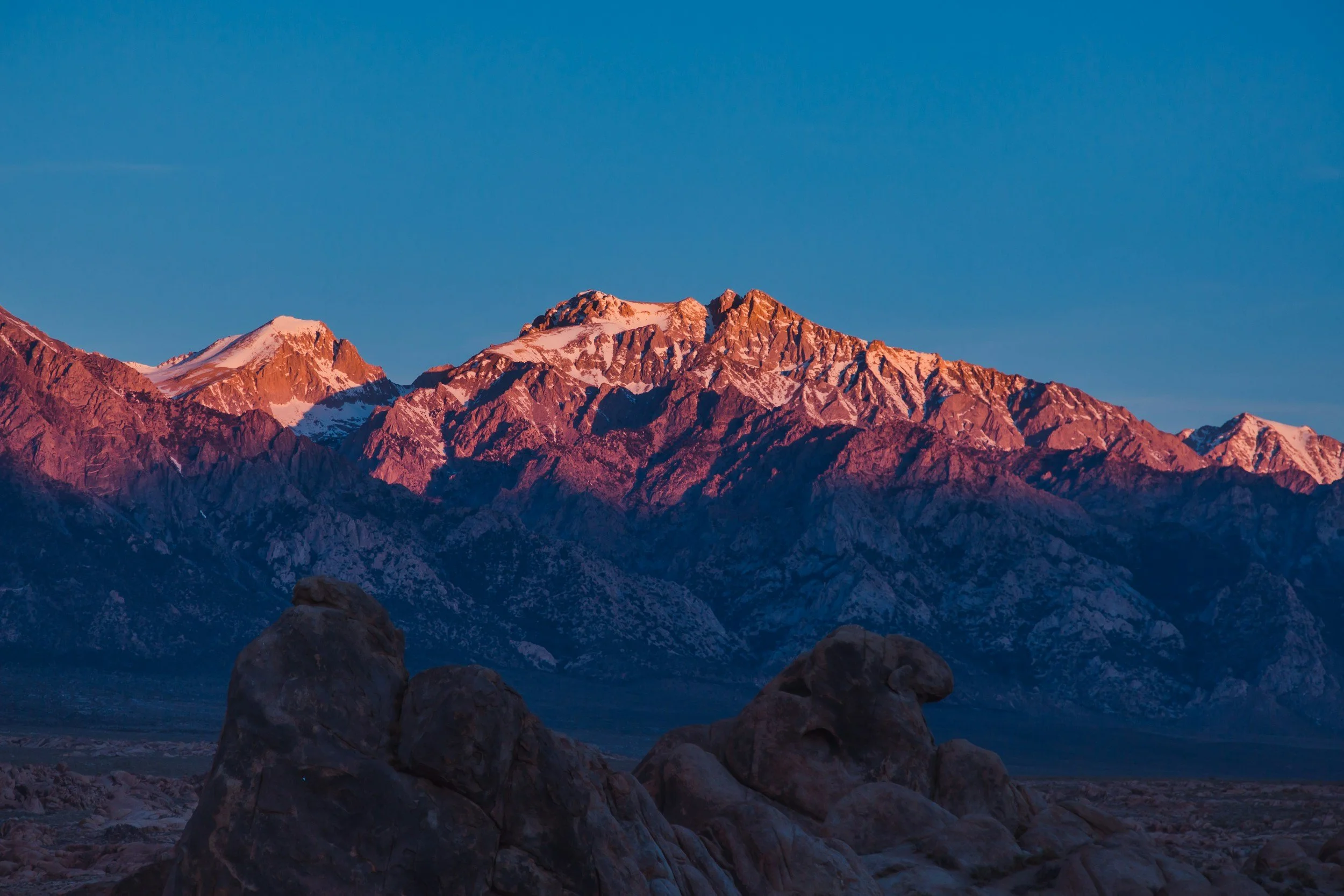 Sunlit mountain peaks with snowcaps and a clear blue sky above, viewed from rocky foreground.