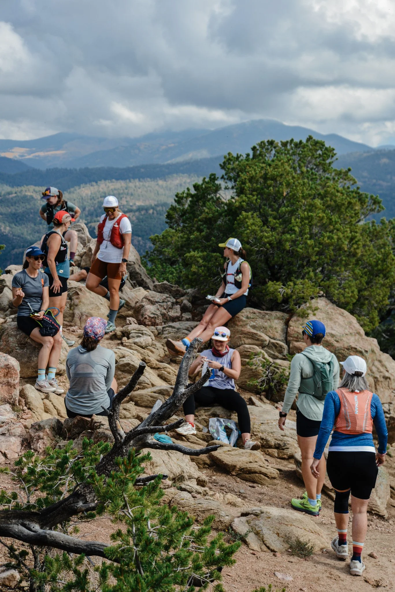 Group of hikers on rocky trail in a mountainous landscape with cloudy sky and evergreen trees.