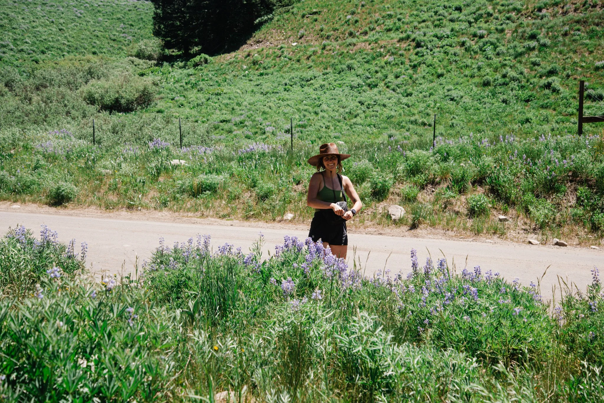 A woman with a large sun hat and sunglasses smiling in a green field of blooming purple flowers with a hillside and trees in the background.
