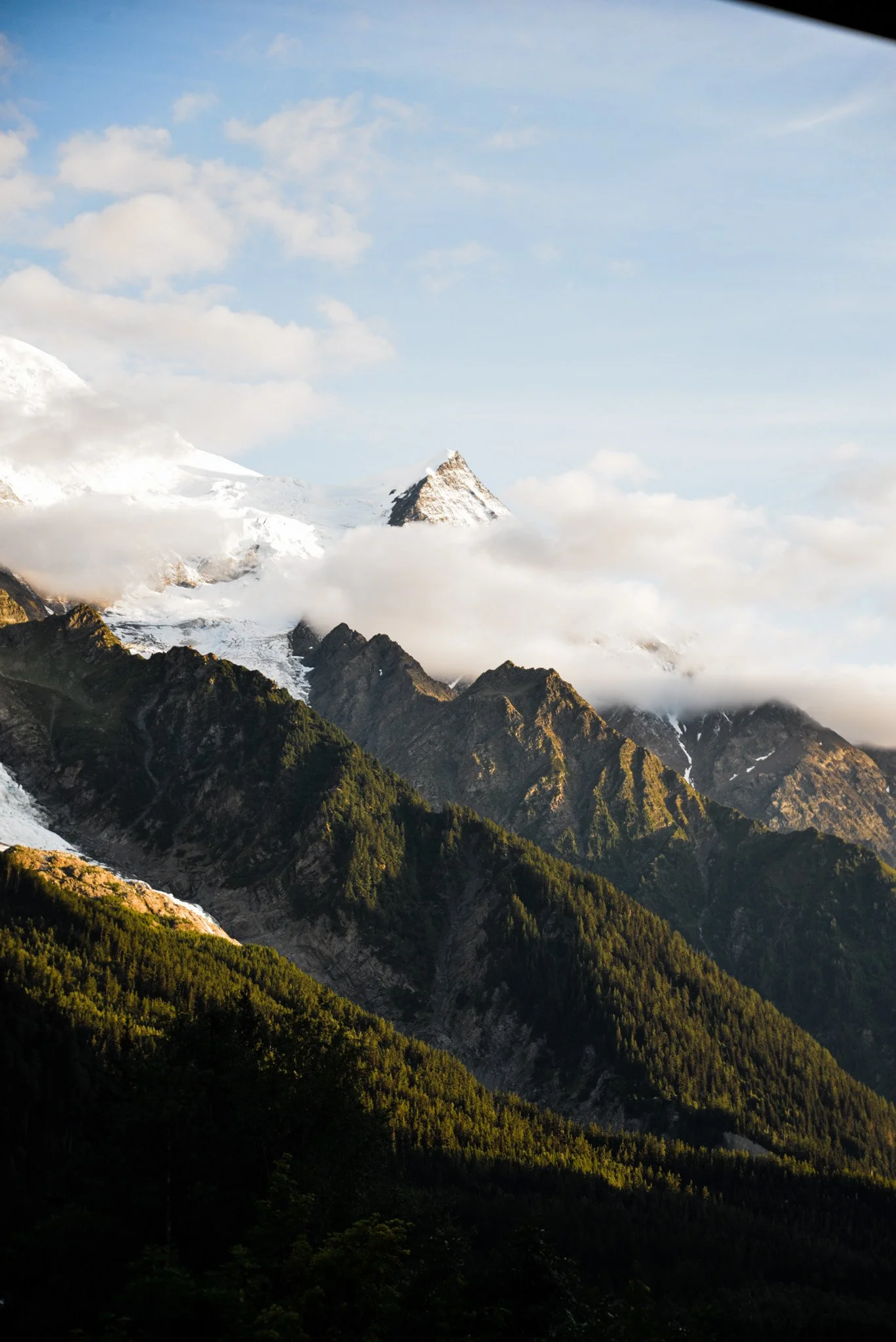 Snow-capped mountain surrounded by clouds, green forested slopes, and a blue sky with scattered clouds.