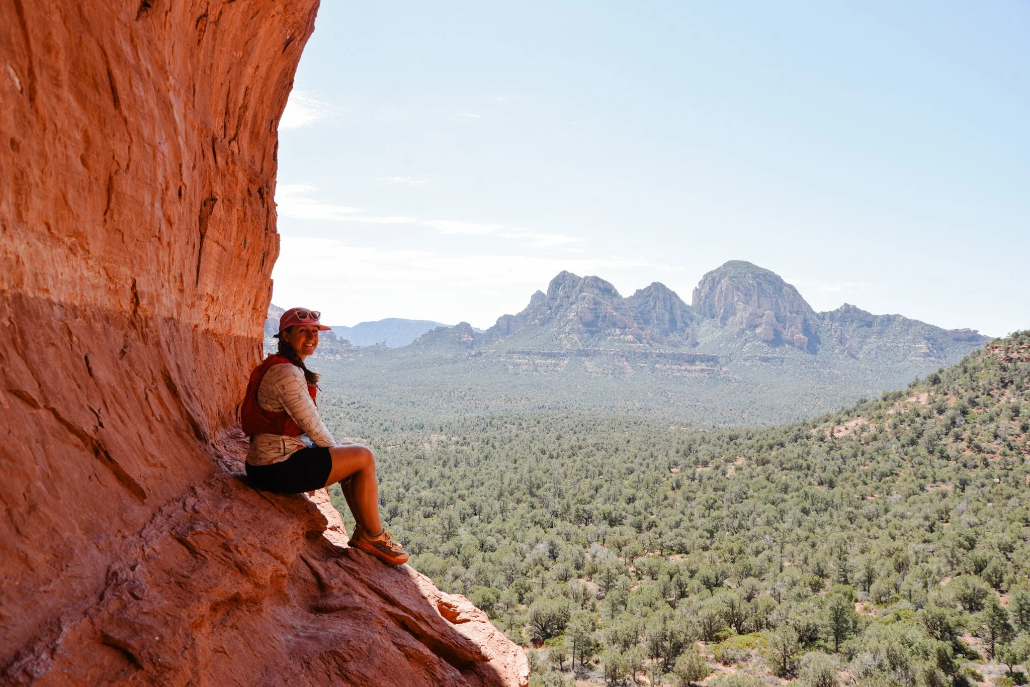 A woman sitting on a red rock cliff with mountains in the background during daytime.