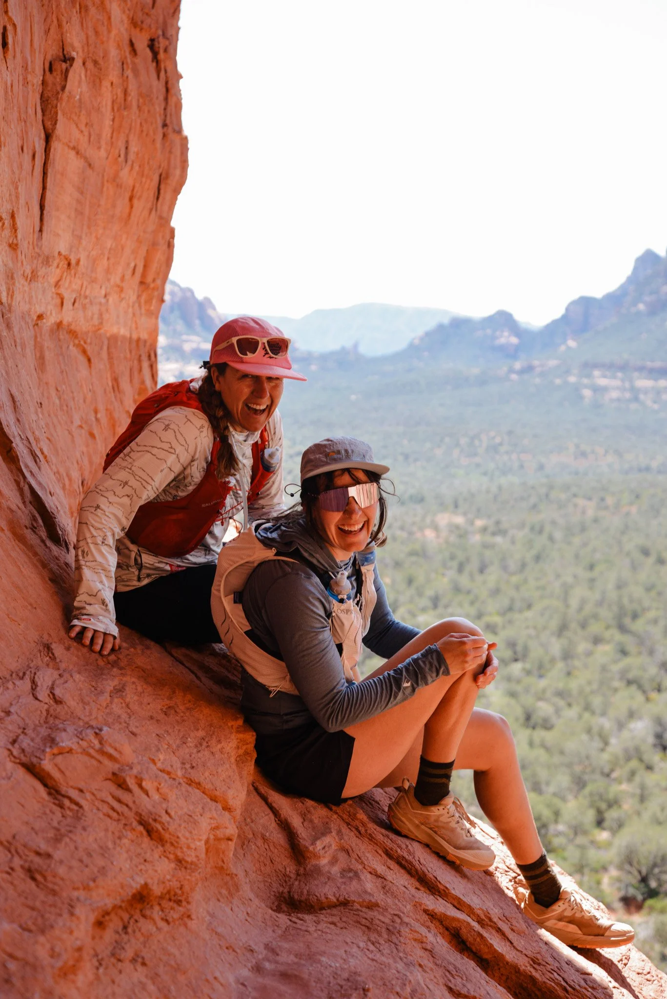 Two women rock climbing on a red cliff, smiling, with a scenic mountain and valley view in the background.