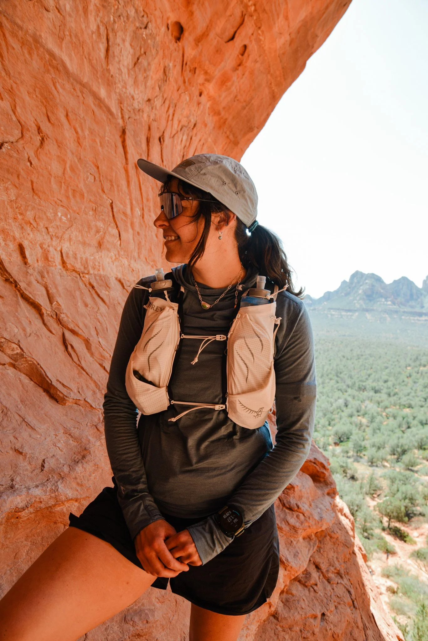 A woman smiling and wearing sunglasses, a visor cap, and outdoor gear, standing next to a red rock formation with scenic mountains and green landscape in the background.