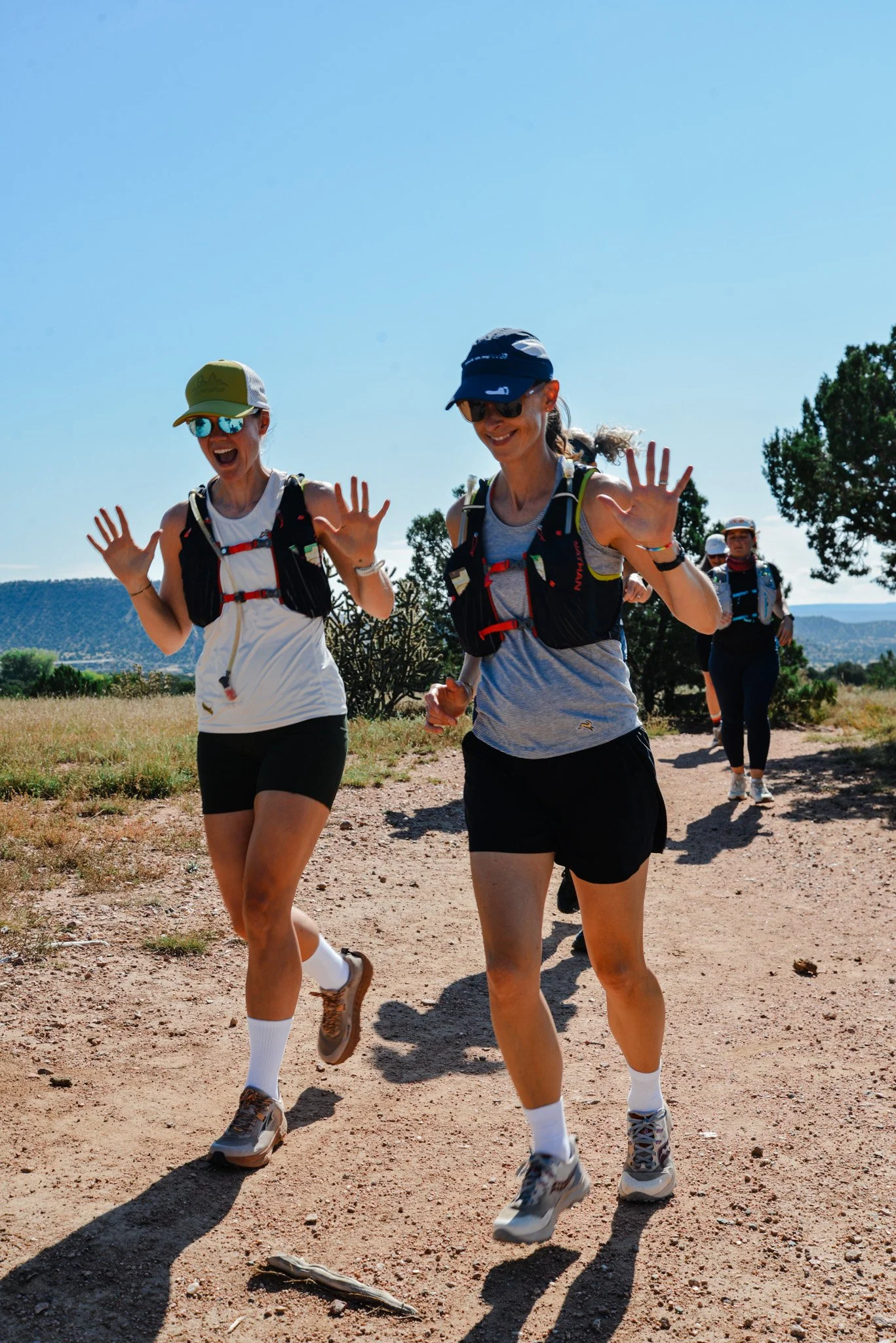 Two women running on a dirt trail outdoors on a sunny day, wearing athletic clothing, sunglasses, caps, and hydration backpacks, with a group of other runners in the background and trees on the side.