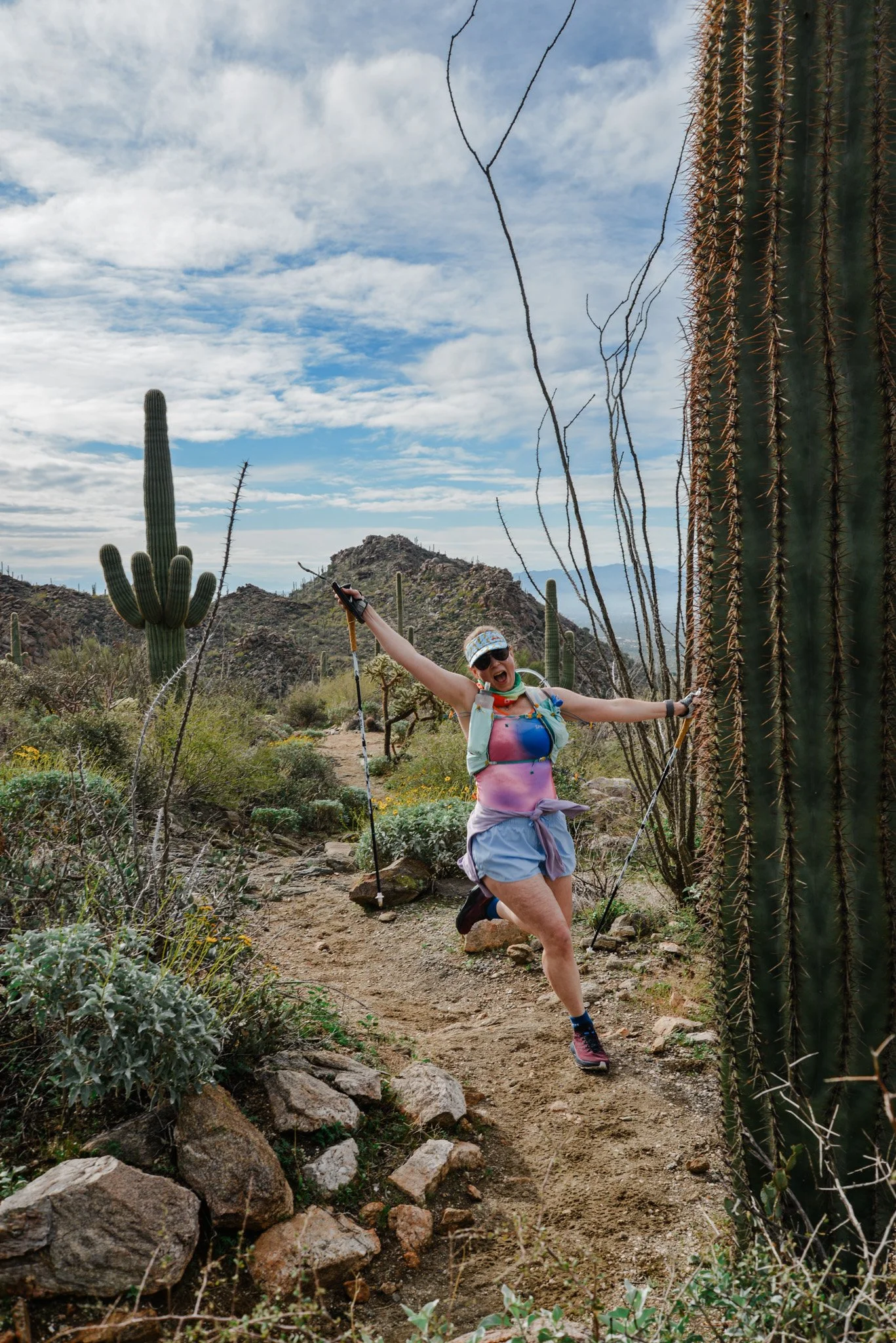 A woman trail running on a rocky dirt path in a desert with saguaro cacti and mountain in the background.