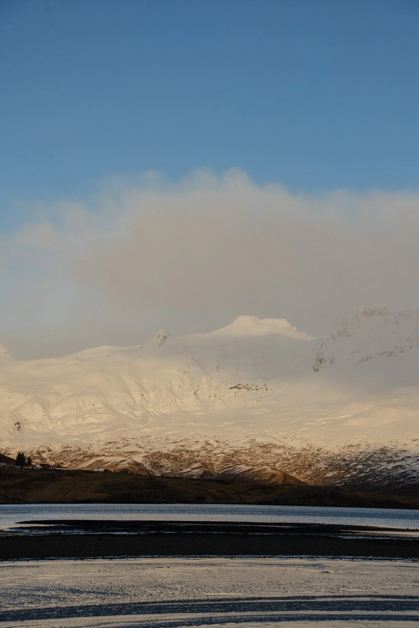 Snow-covered mountain landscape with a body of water in the foreground and a small settlement at the base of the mountains.