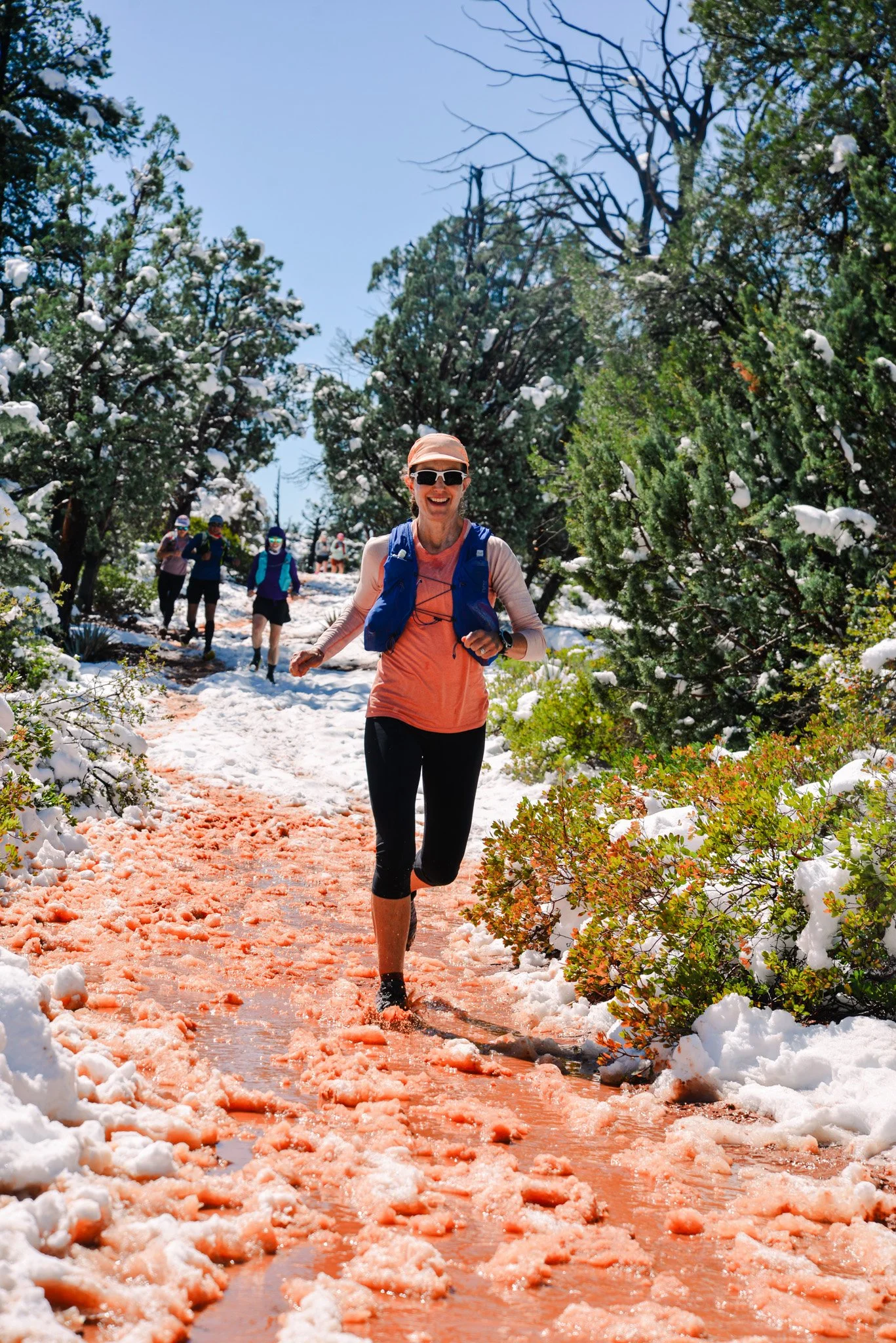A woman jogging on a snowy trail with snow-covered trees around her, a group of trail runners in the background, sunny weather, and a clear blue sky.