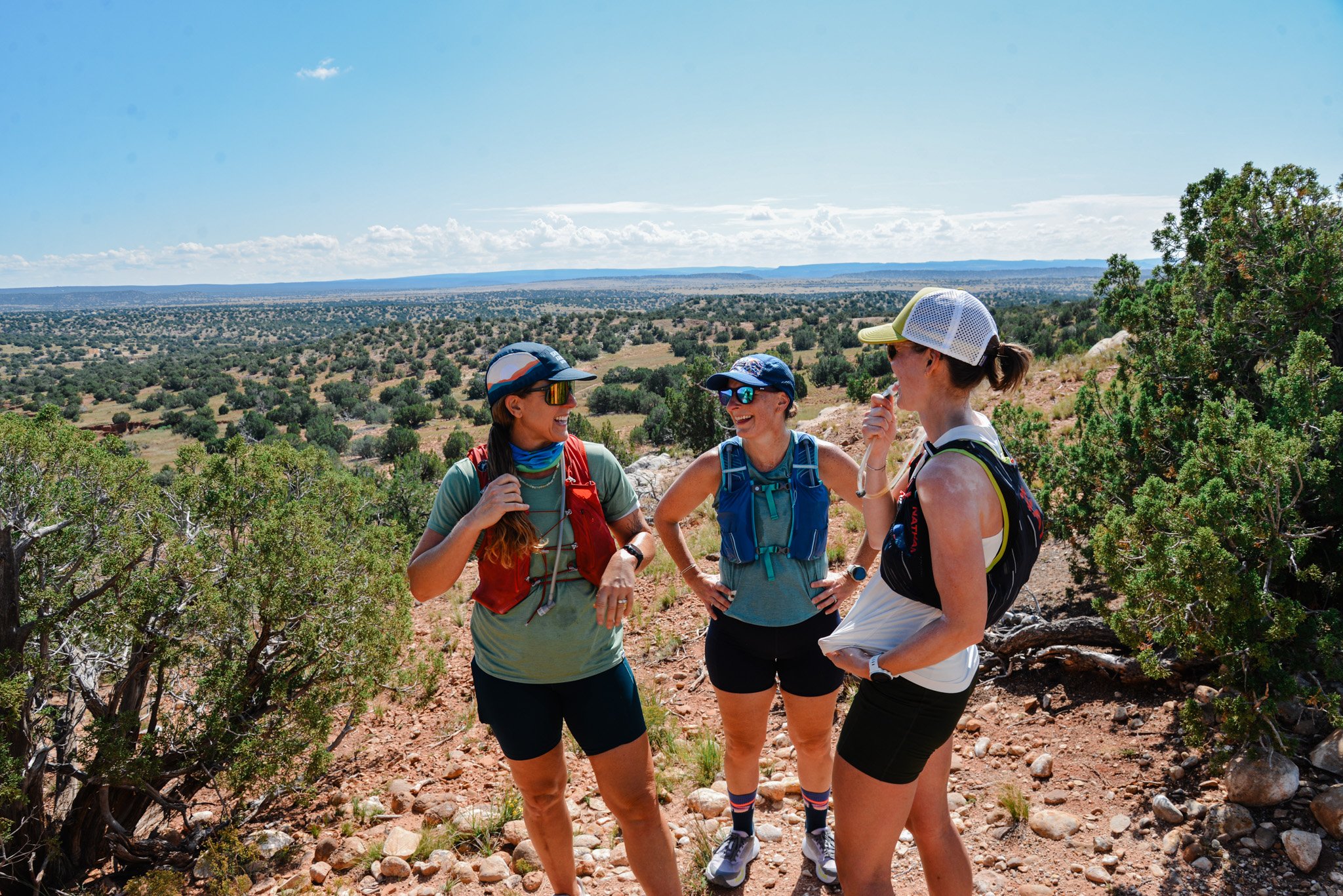 Four women hikers standing together and smiling outdoors in a desert landscape, wearing casual hiking gear, hats, and sunglasses, engaging in conversation.