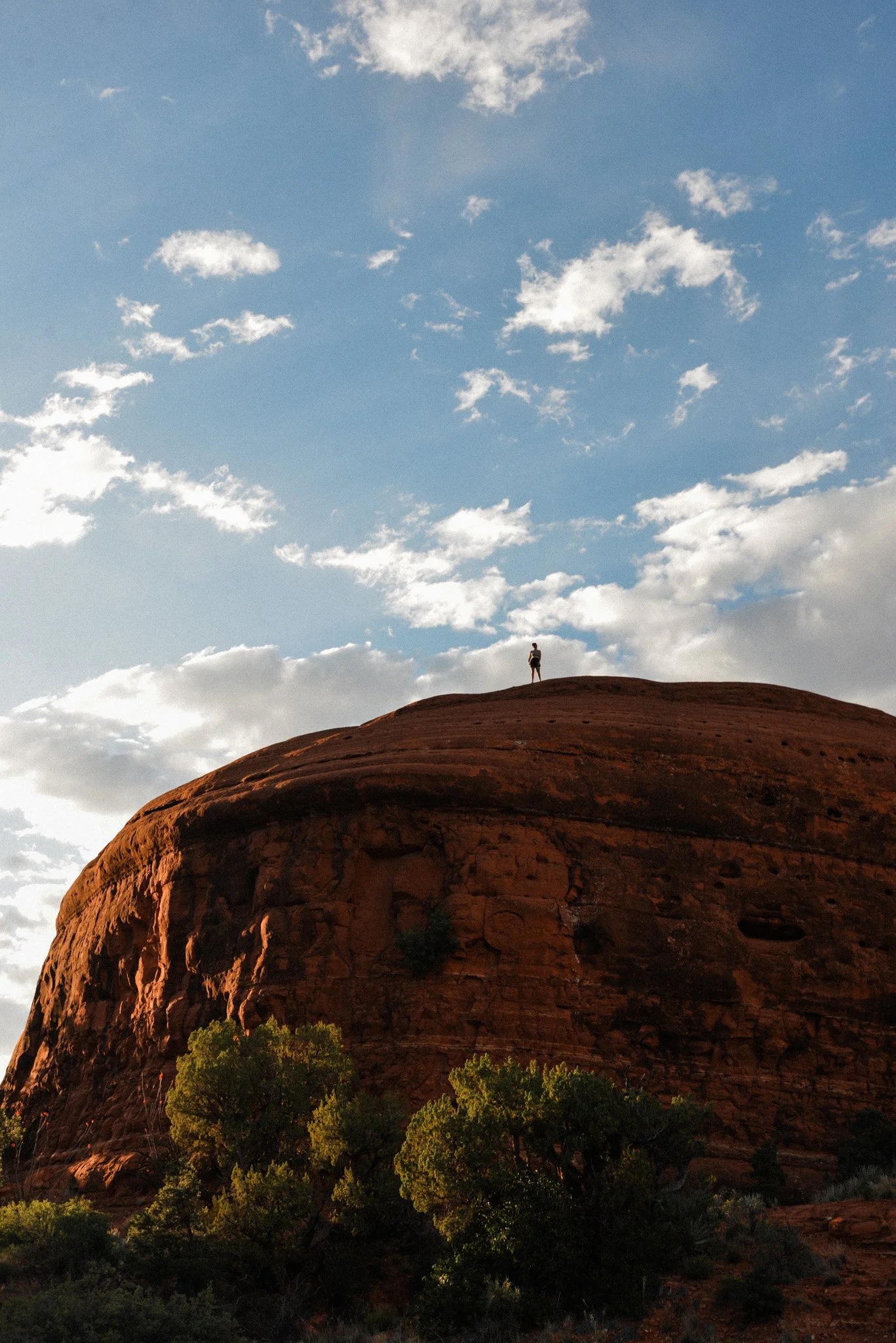 A person standing on top of a large red rock formation, with a sky filled with scattered clouds in the background and green trees at the base.