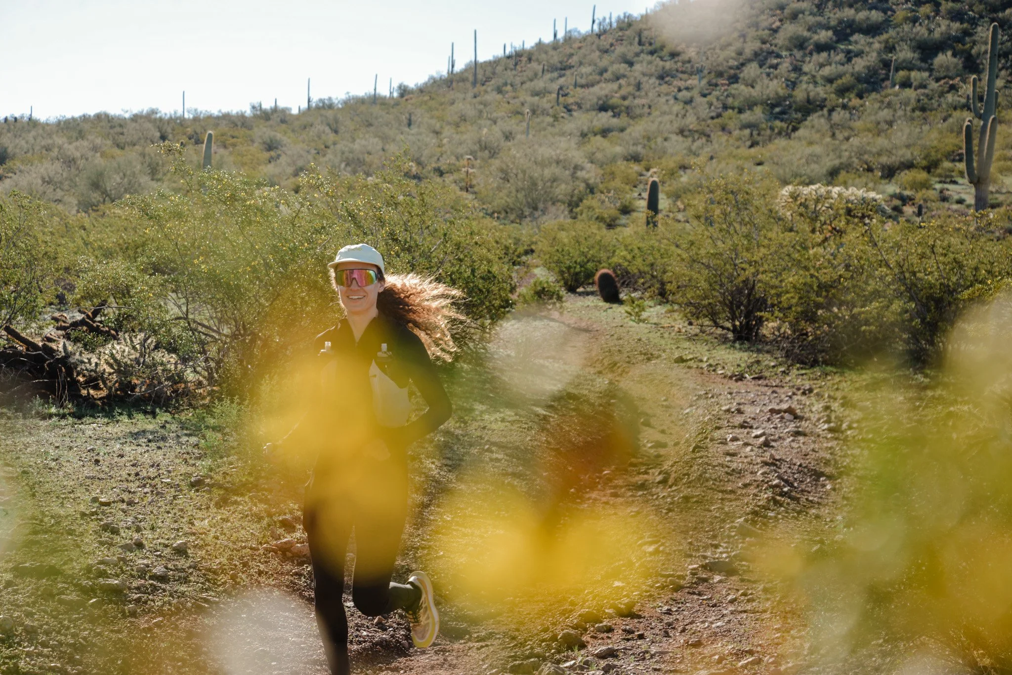 A woman running on a dirt trail in a desert landscape with numerous saguaro cacti and sparse vegetation under a clear sky.