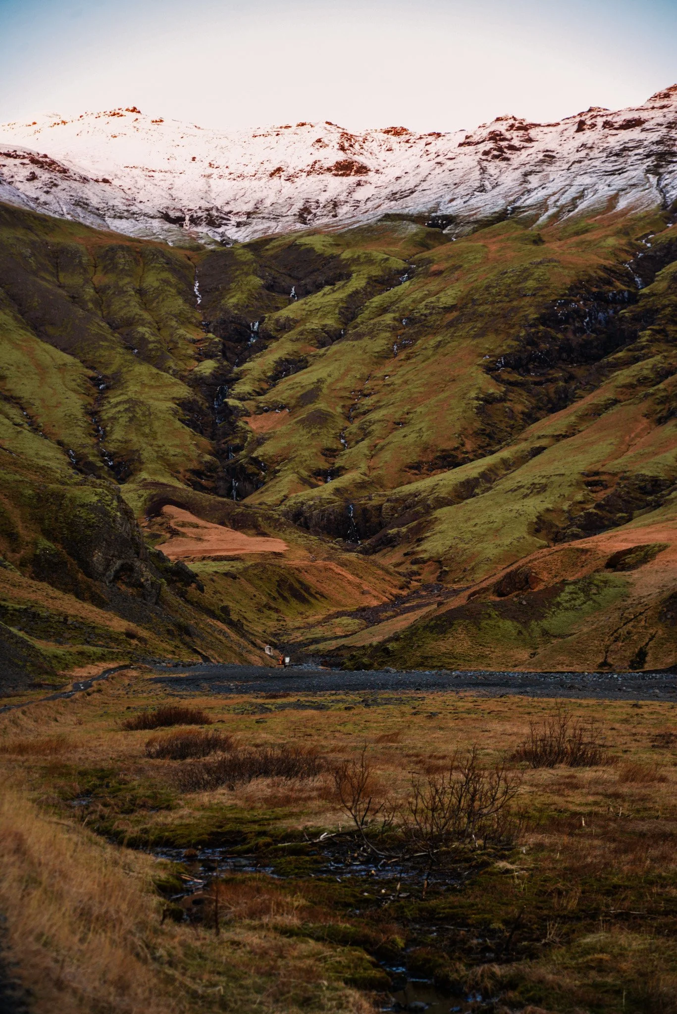 A mountain landscape with snow-capped peaks, green and brown sloped hills, small streams, and a flat grassy area in the foreground.