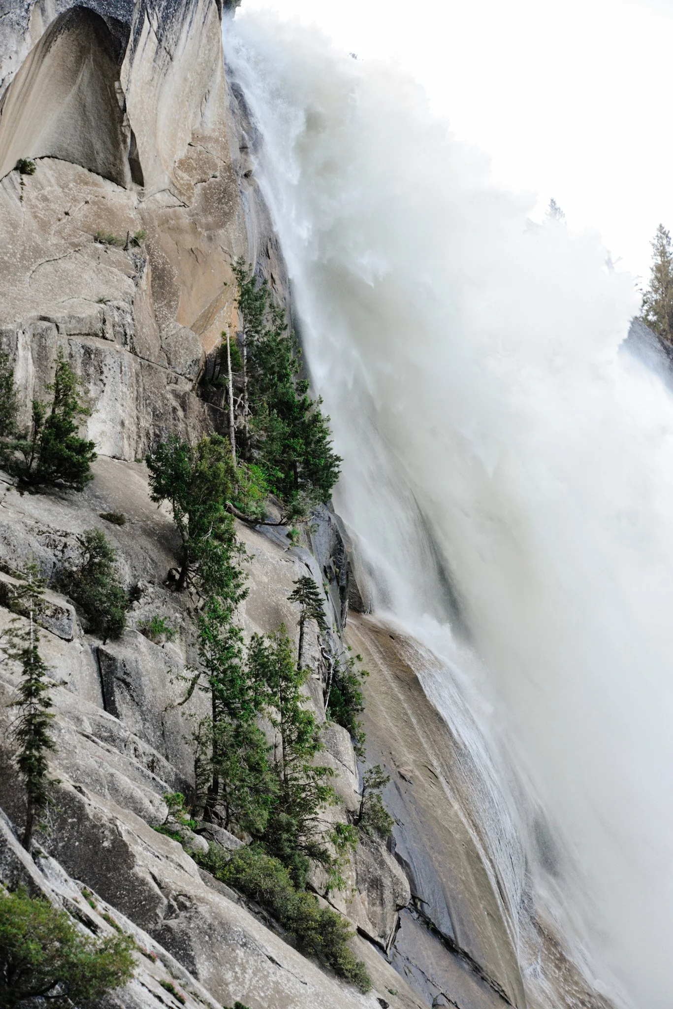 View of a waterfall with water crashing down a rocky cliff face, surrounded by green pine trees.