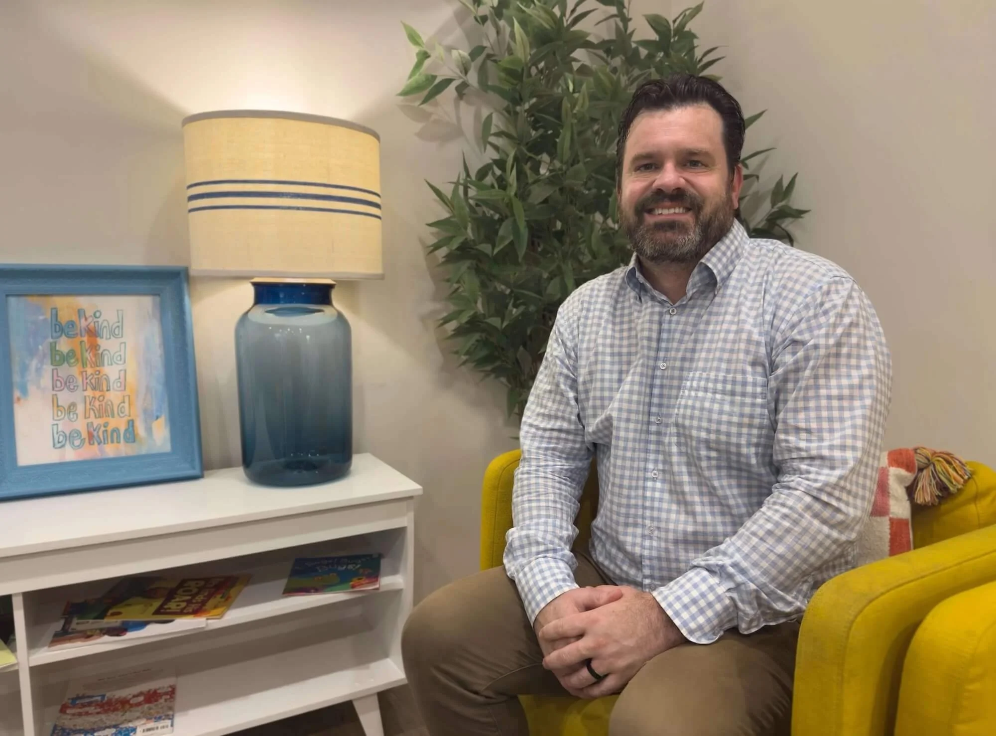 Man with dark hair and beard smiling, wearing a light checkered shirt, sitting in a room with a yellow chair, beside a white shelf with children's books and a framed artwork that says "be kind." Behind him, a plant and a beige wall with a lamp.