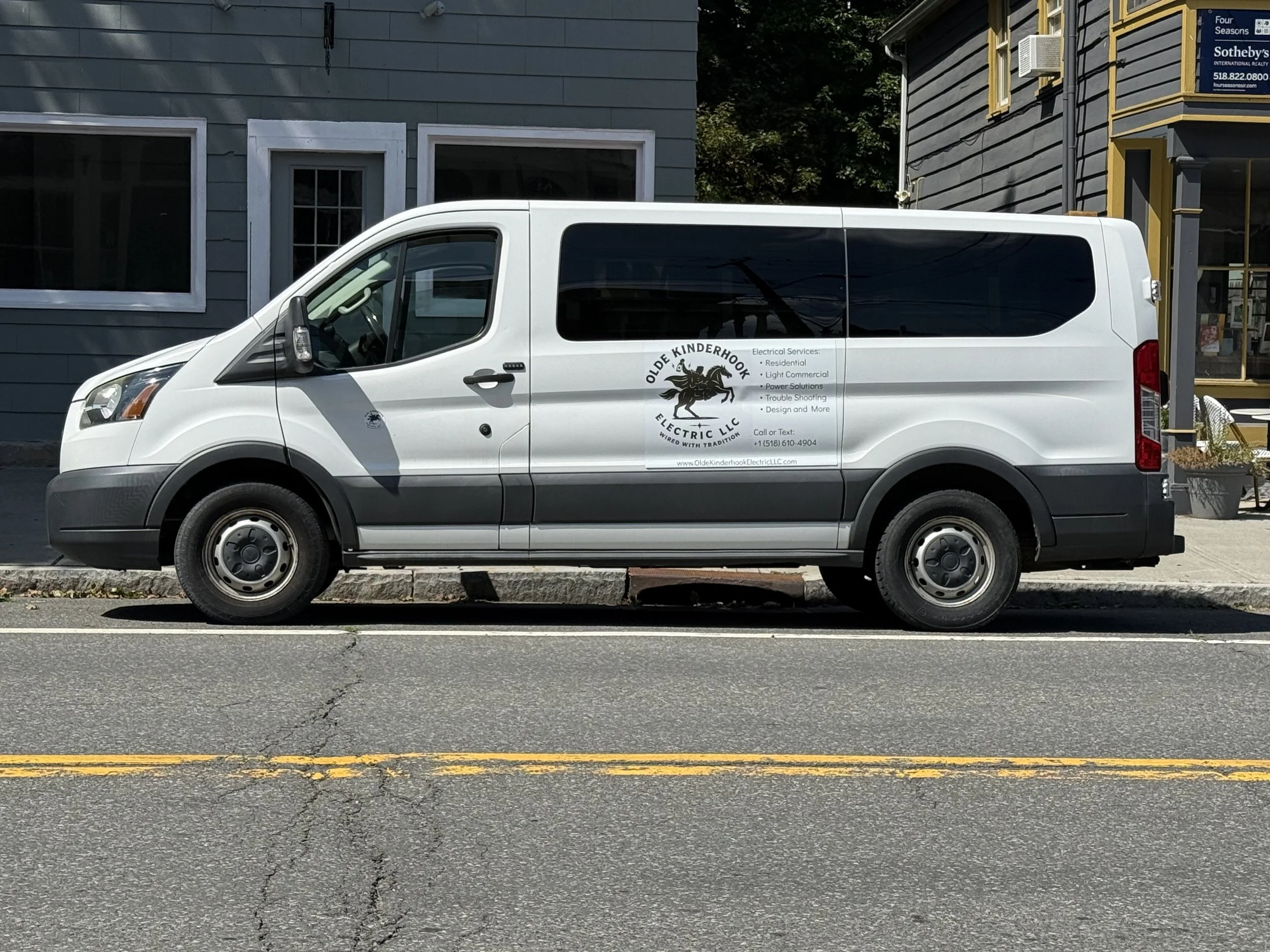 White van parked on the street in front of gray and yellow buildings with a sign that reads 'Olde Kinderhook Electric LLC'.