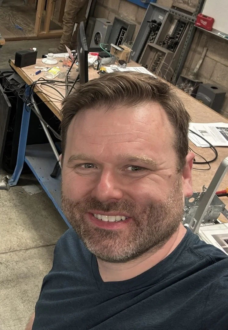 A man taking a selfie in a workshop or industrial setting with workbenches, tools, and equipment in the background.
