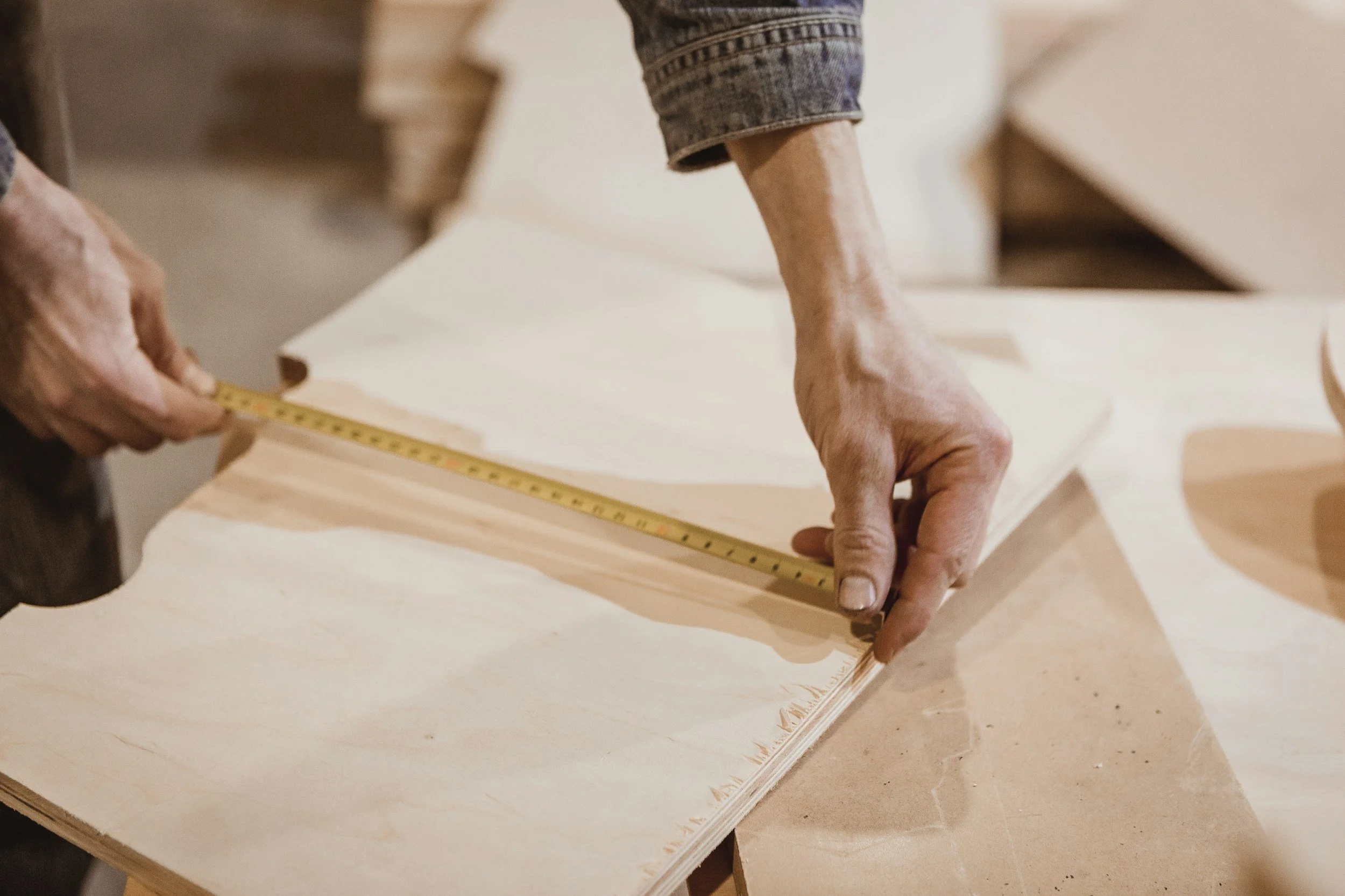 An elderly hand measuring a piece of light-colored wood with a yellow tape measure at a woodworking or carpentry workbench.