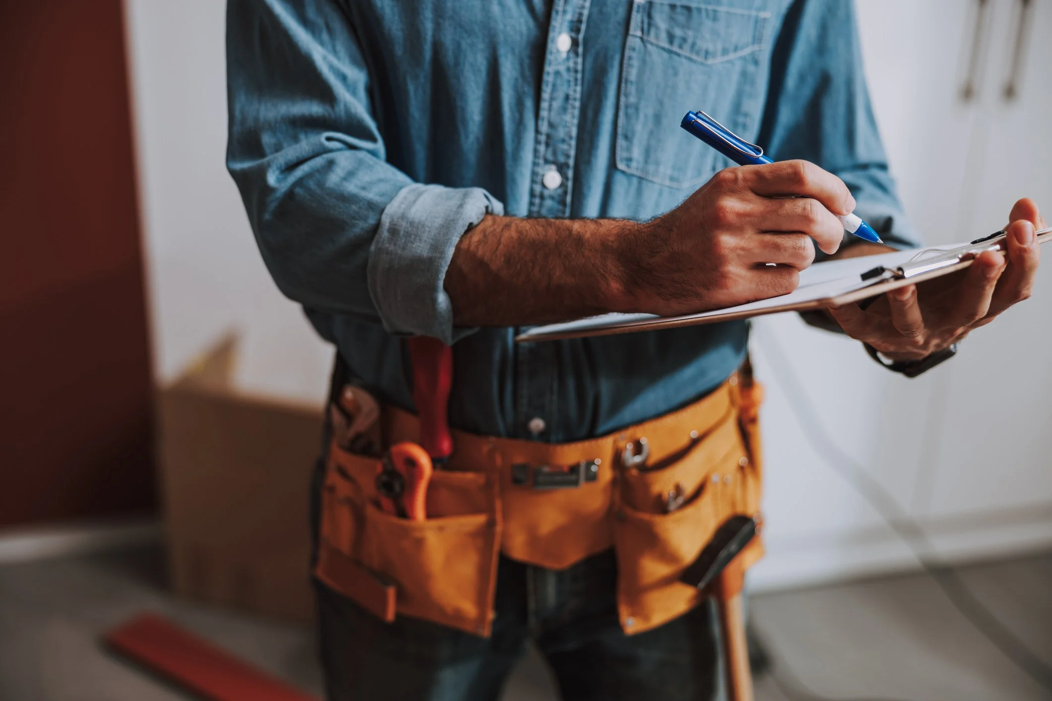 Close-up of a person wearing a denim shirt and orange work pants, holding a clipboard and a pen, with tools attached to the pants belt.