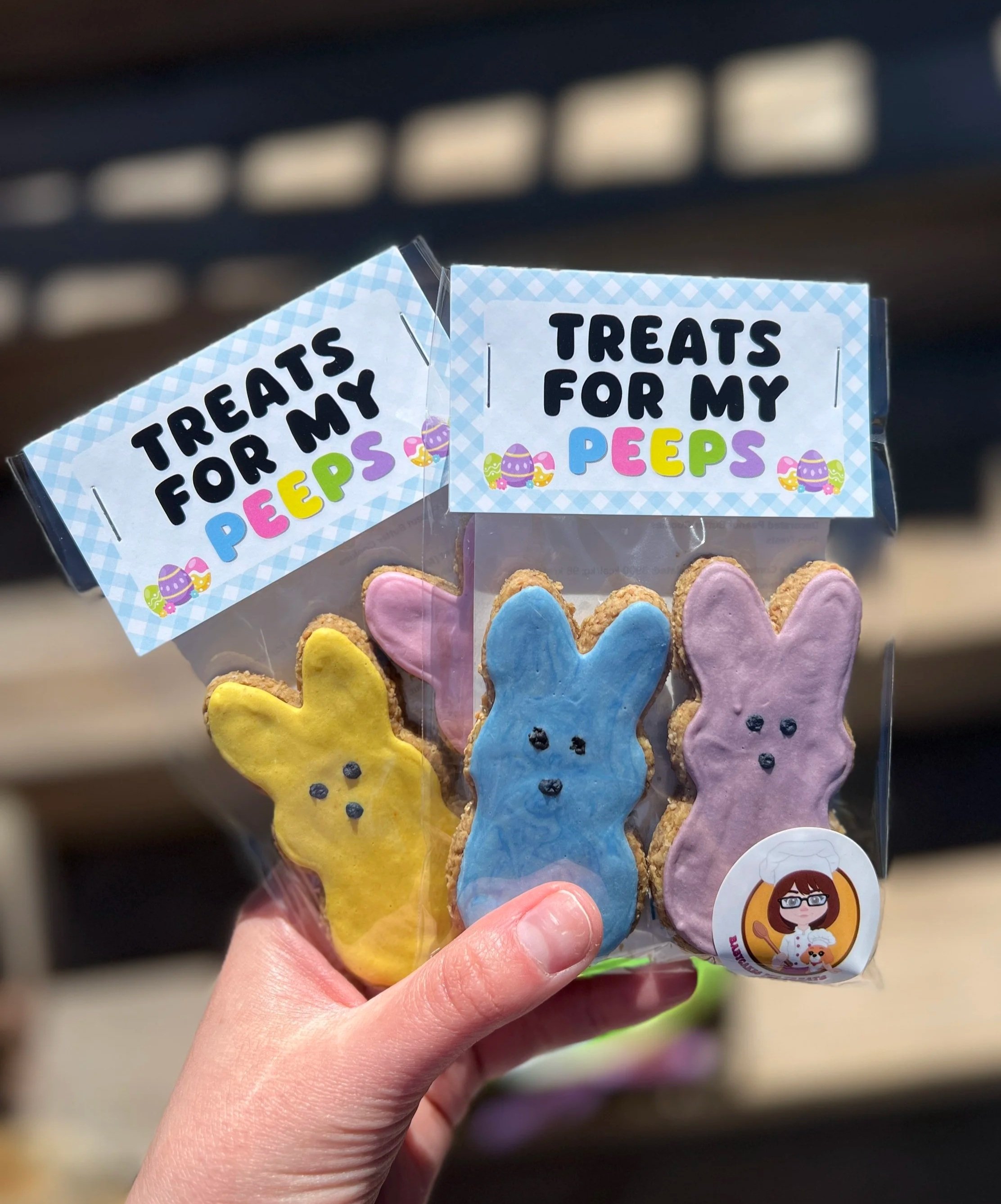 Hand holding a clear plastic bag with decorated bunny-shaped cookies in yellow, pink, blue, and purple, with a spring-themed tag featuring Easter eggs for dogs and a cartoon girl wearing glasses and a chef's hat.