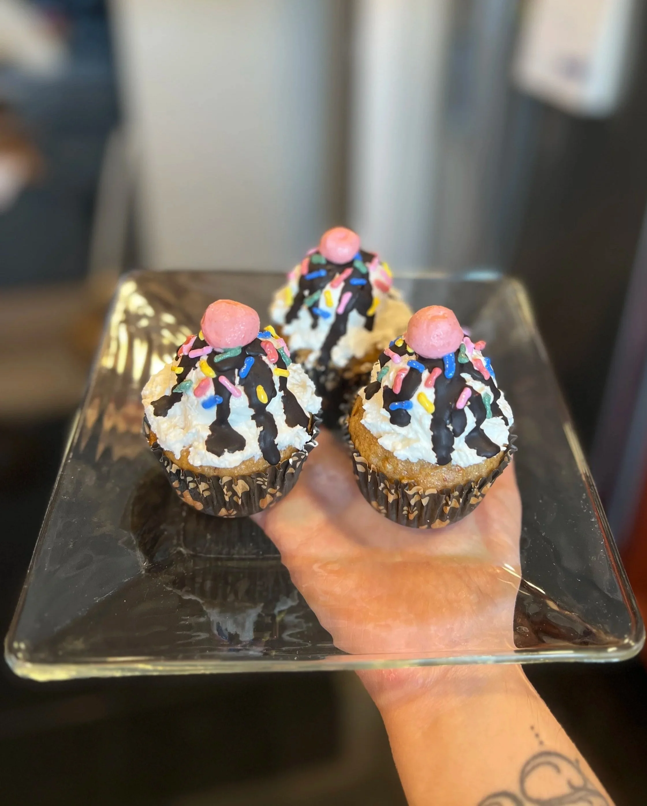 Close-up of a tray holding three cupcakes decorated with pink, rainbow, and black sprinkles, topped with pink candies, for dogs, held by a person with a tattooed arm.