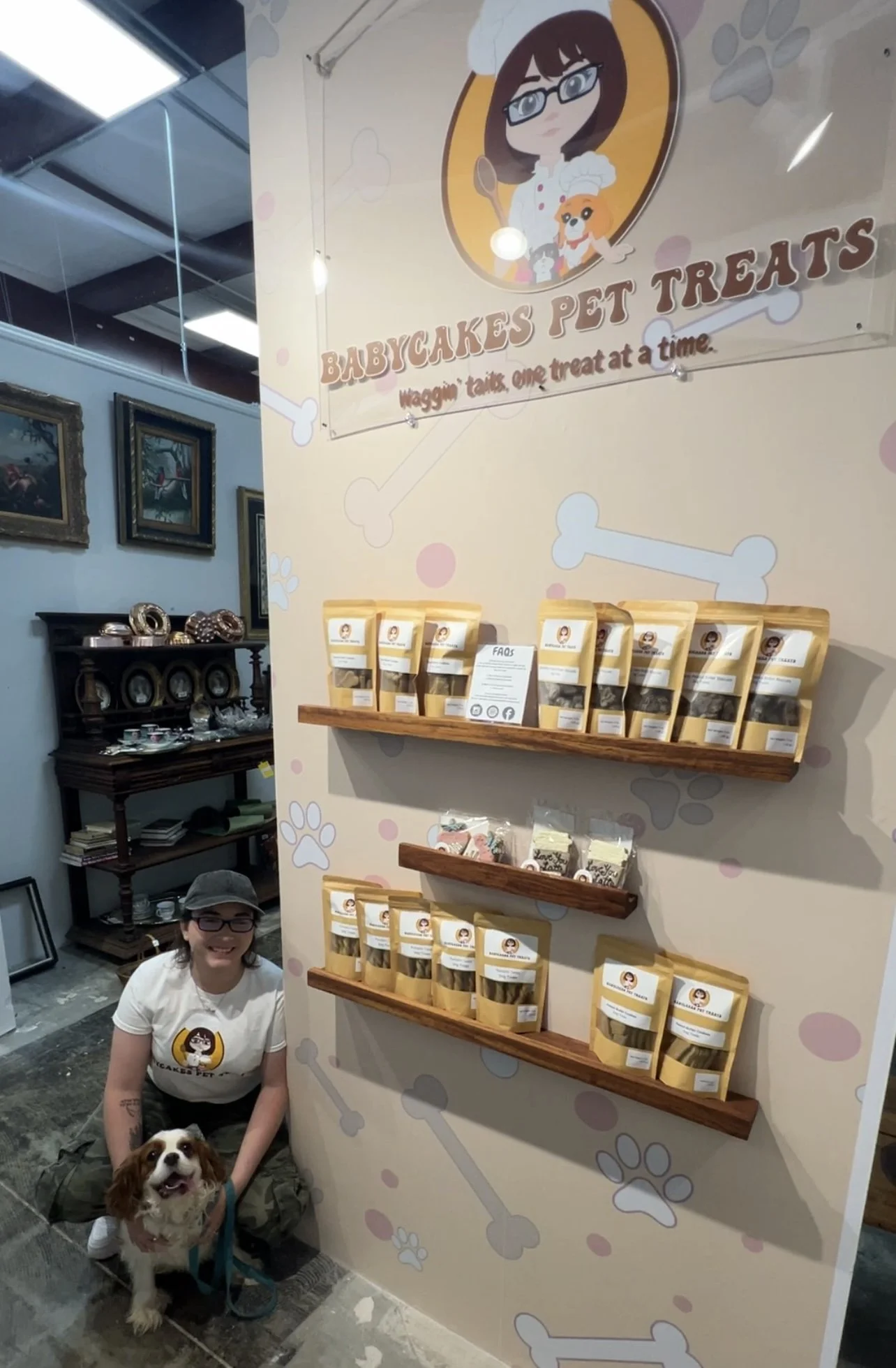 A woman with glasses and a cap, kneeling next to a dog, is posing in front of a display wall at a pet treat booth. The wall has branding for Babycakes Pet Treats with an illustration of a female chef dog character, shelves with packaged treats, and a person in the background inside an art gallery or retail space.