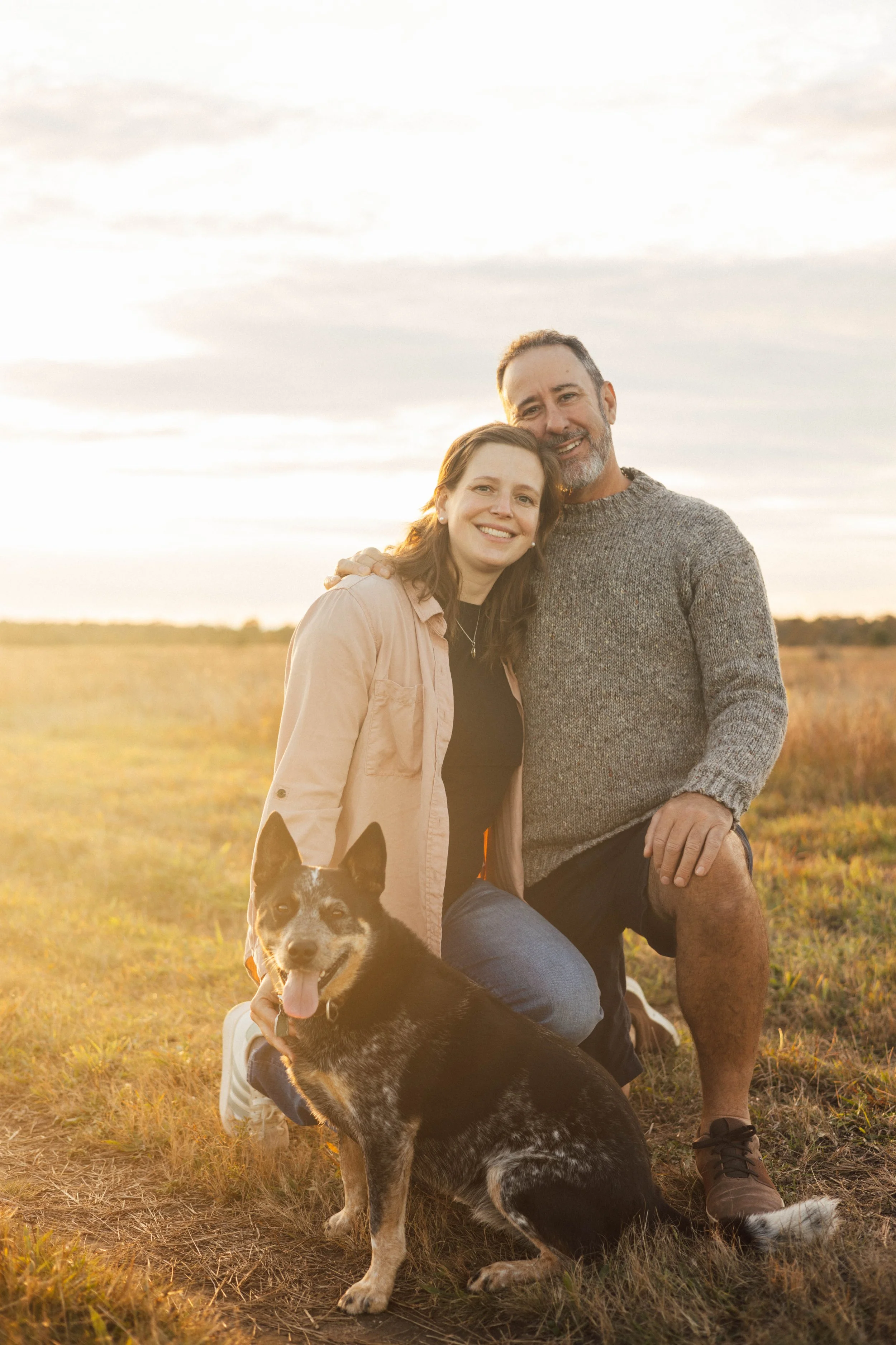 A smiling woman and man posing outdoors in a field at sunset with a black and white dog sitting in front of them.