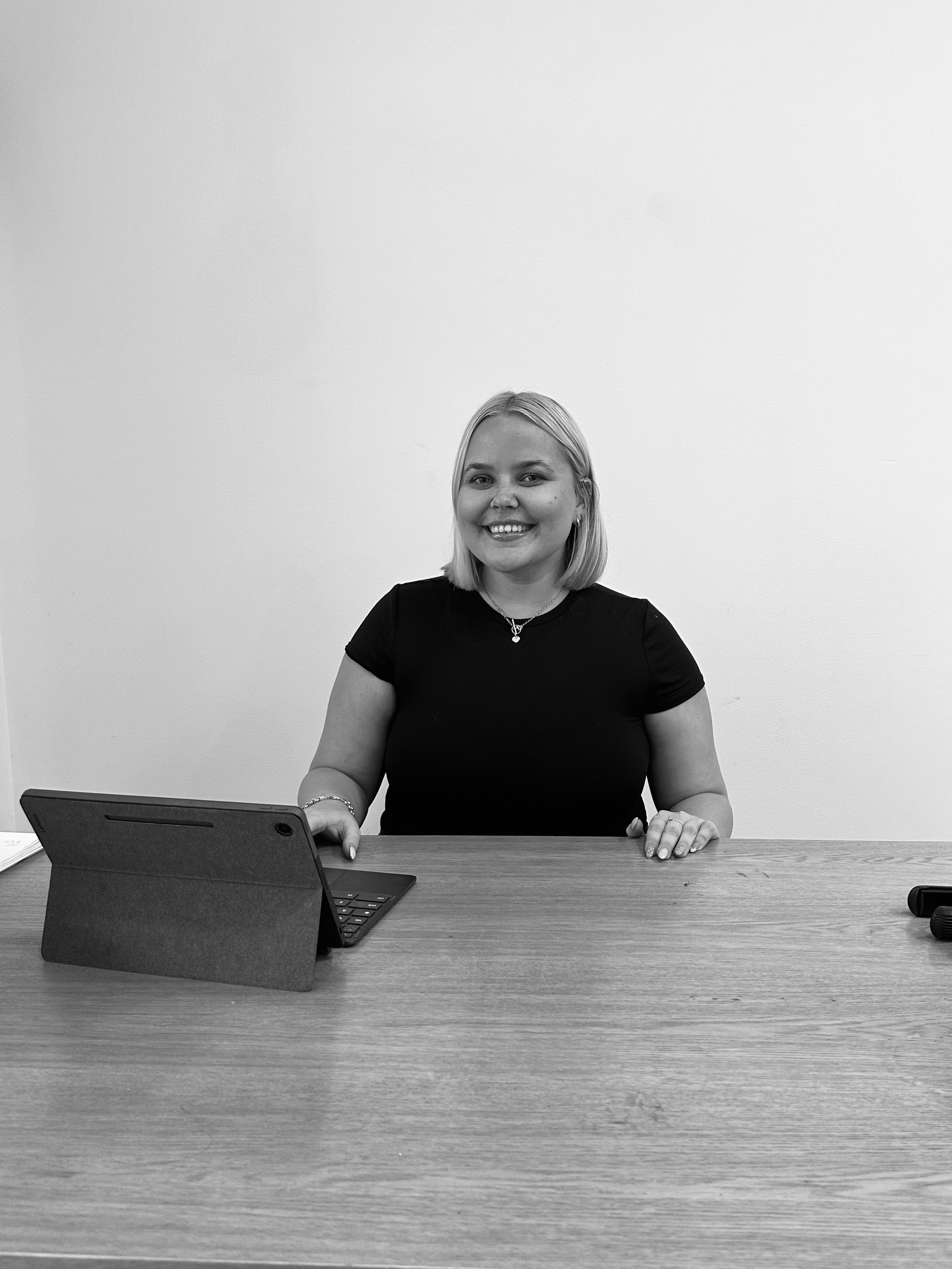 A smiling woman with shoulder-length blonde hair sitting at a desk, wearing a black top and jewelry, with a laptop and smartphone on the desk against a plain white wall.