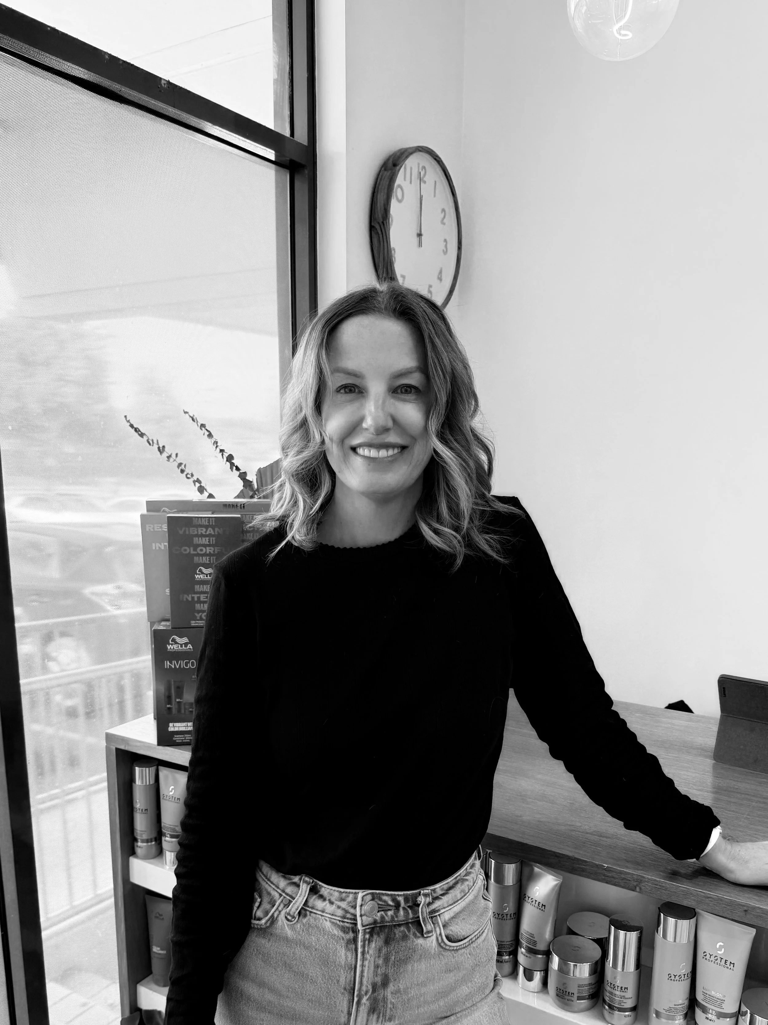 Black and white photo of a woman with wavy hair, smiling, standing behind a counter in a store or salon, with shelves of hair products behind her and a wall clock on the wall.