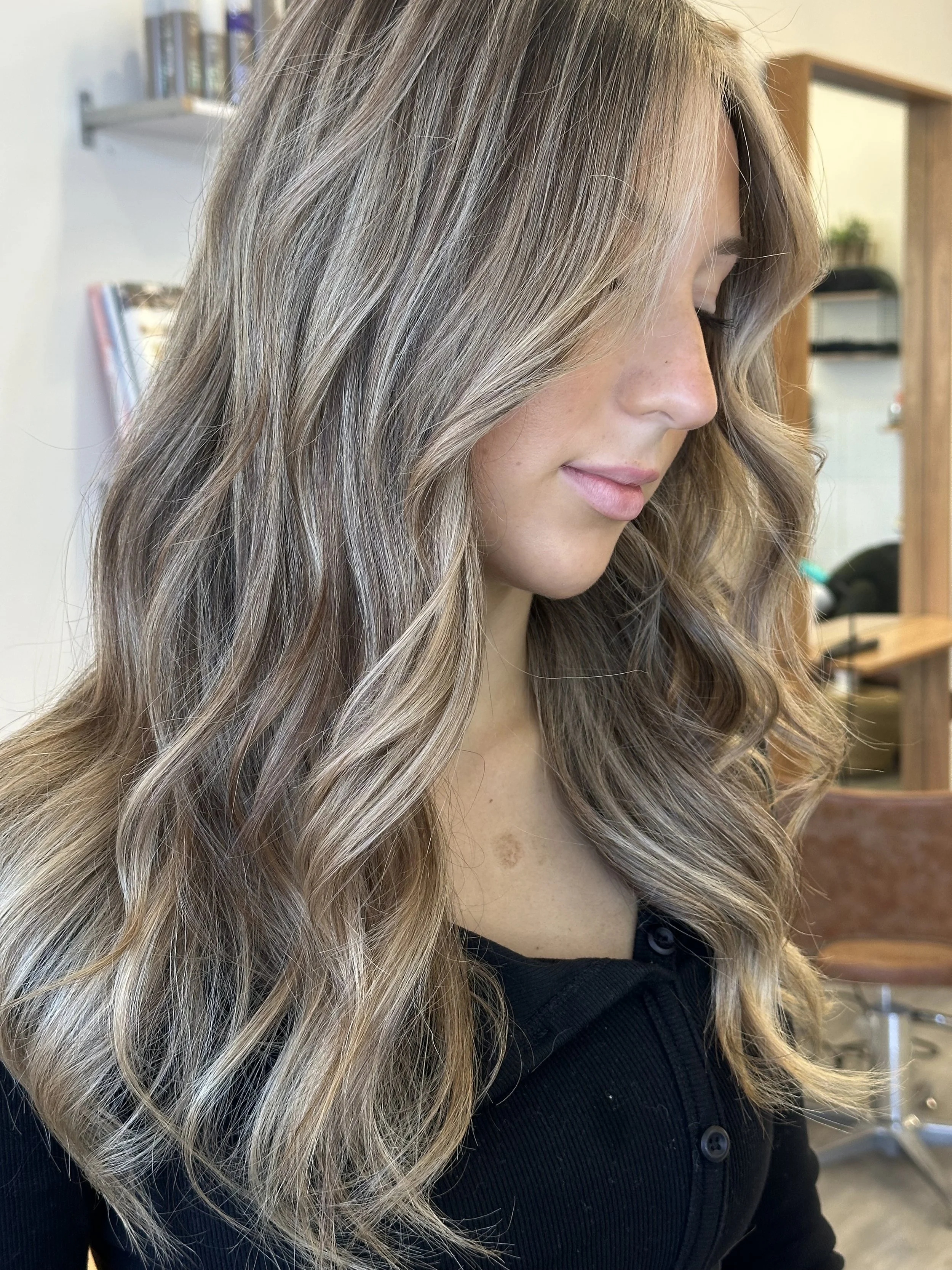 A woman with long, wavy blonde hair with darker roots, wearing a black top, appears to be in a salon or similar setting.