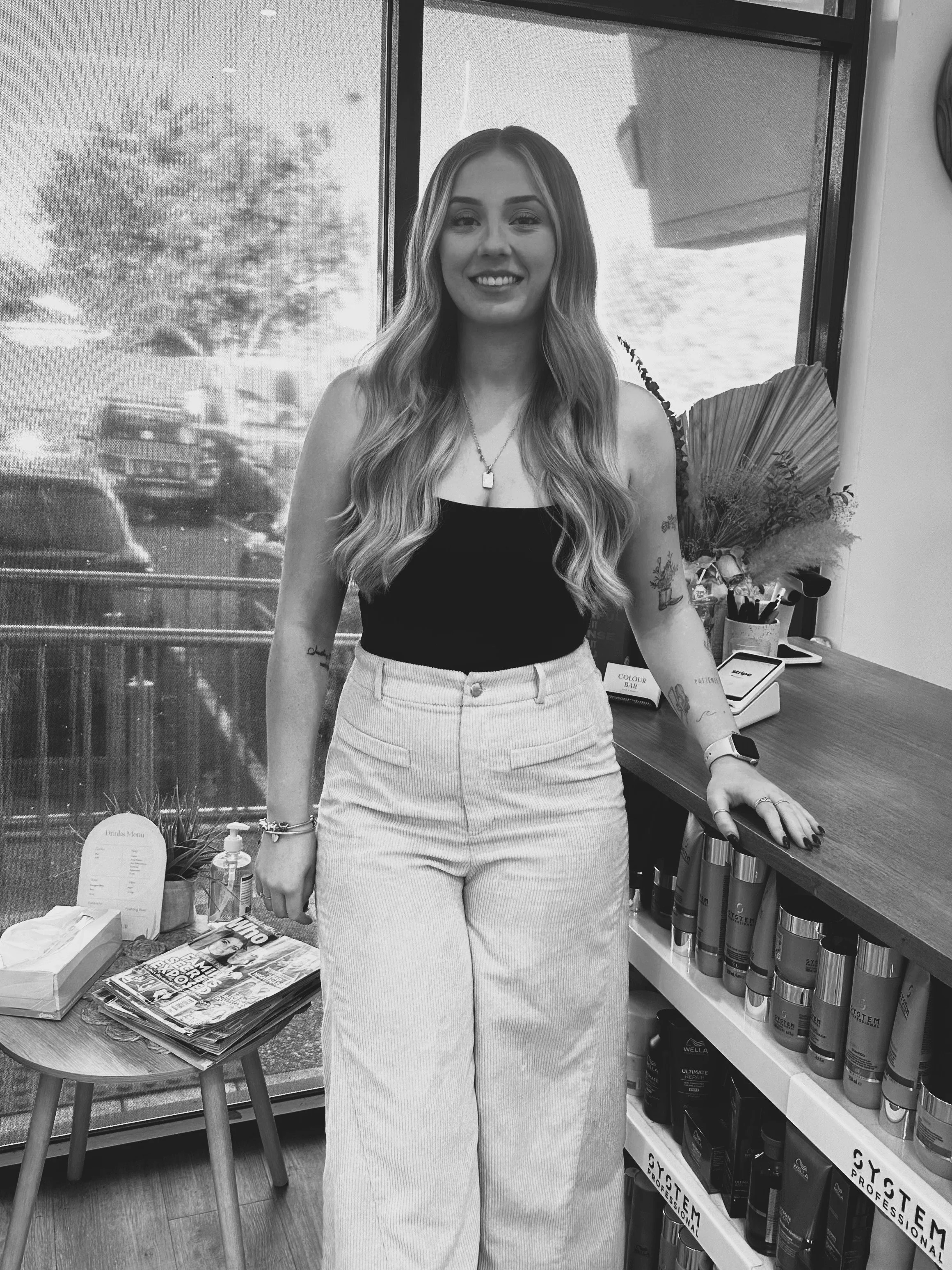 A woman with long wavy hair, smiling, wearing a black top and high-waisted pants, standing inside a store near a window and a shelf with hair products.