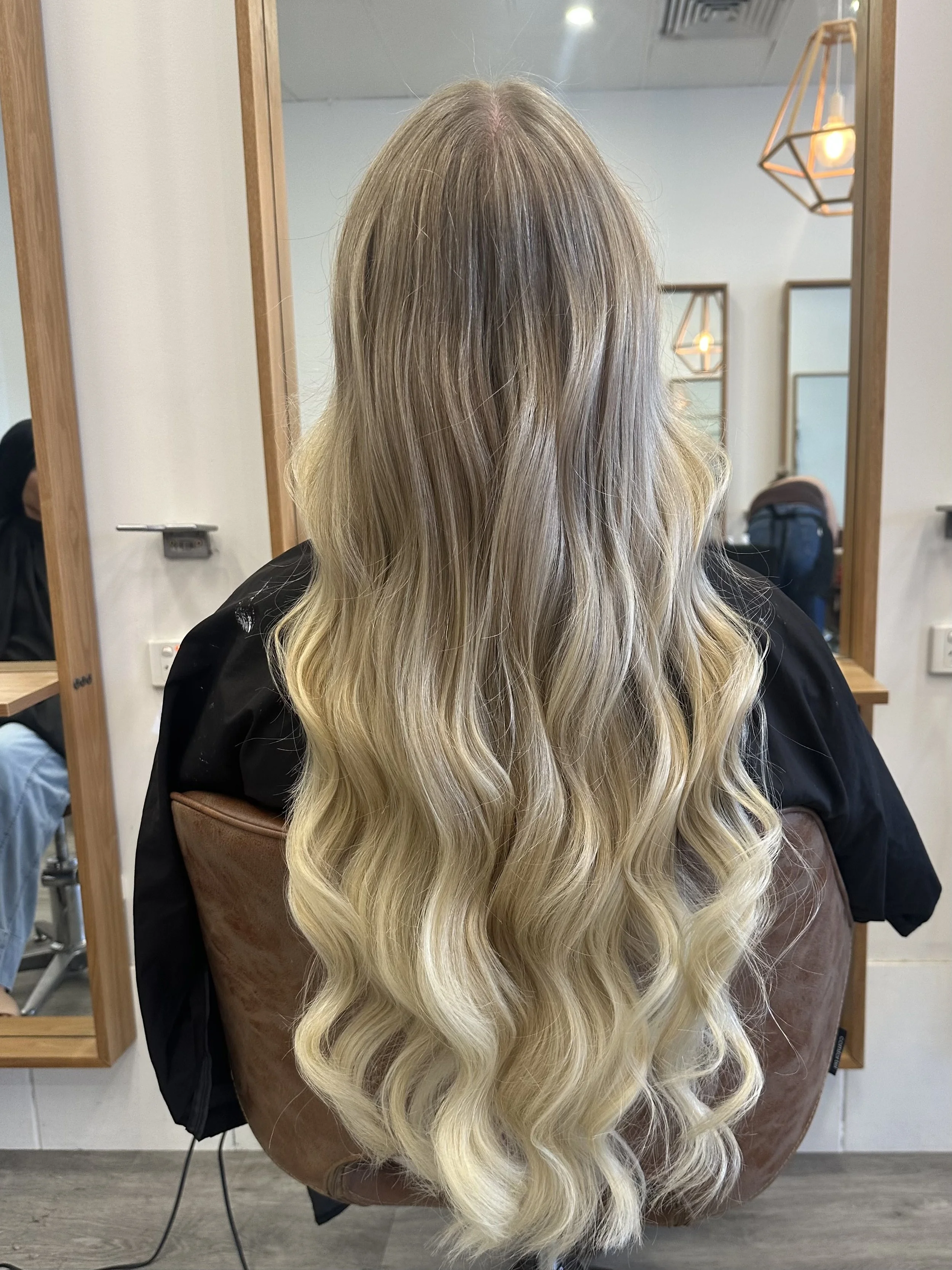 Long blonde wavy hair seen from the back, in a salon setting with mirrors and salon lighting