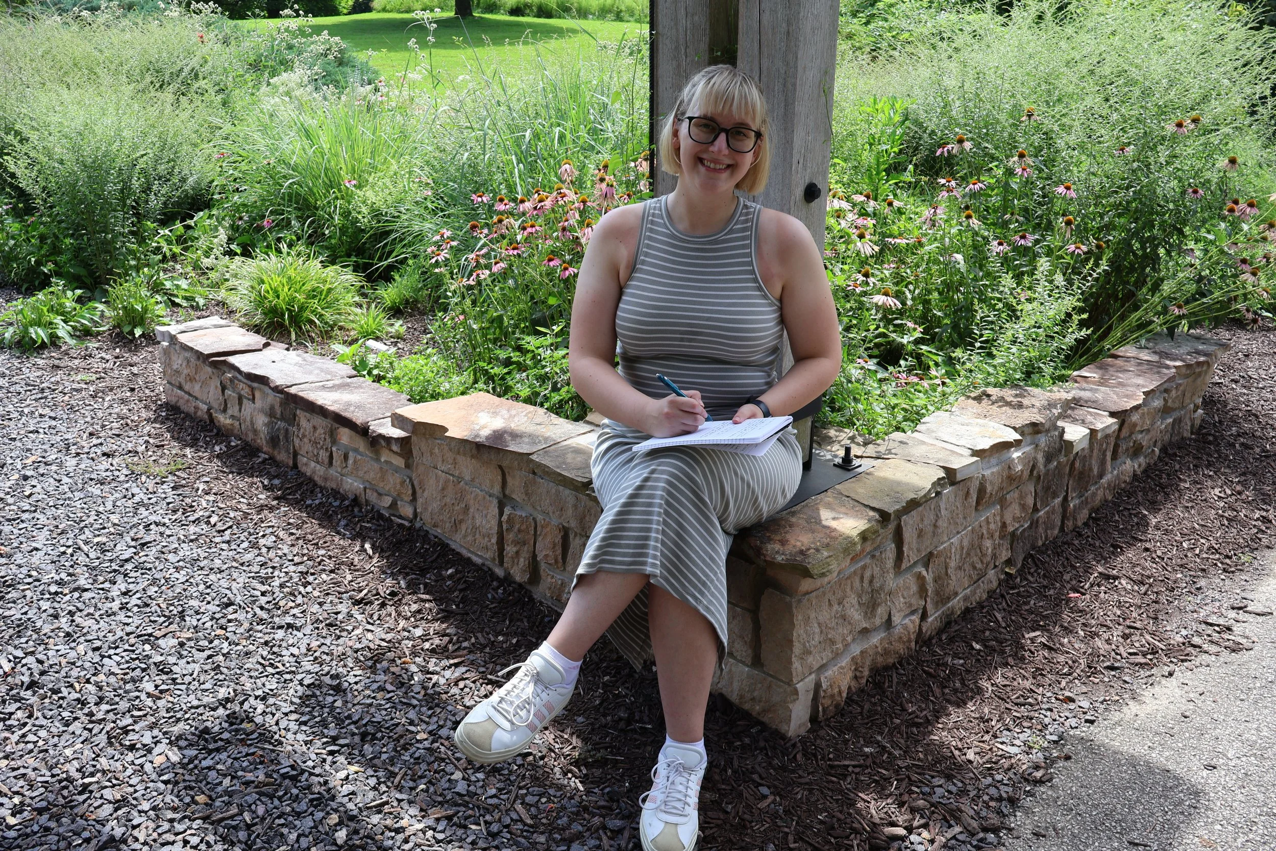 A young woman with short blond hair, glasses, and a striped sleeveless dress sitting on a stone ledge in a garden, smiling and writing in a notebook.