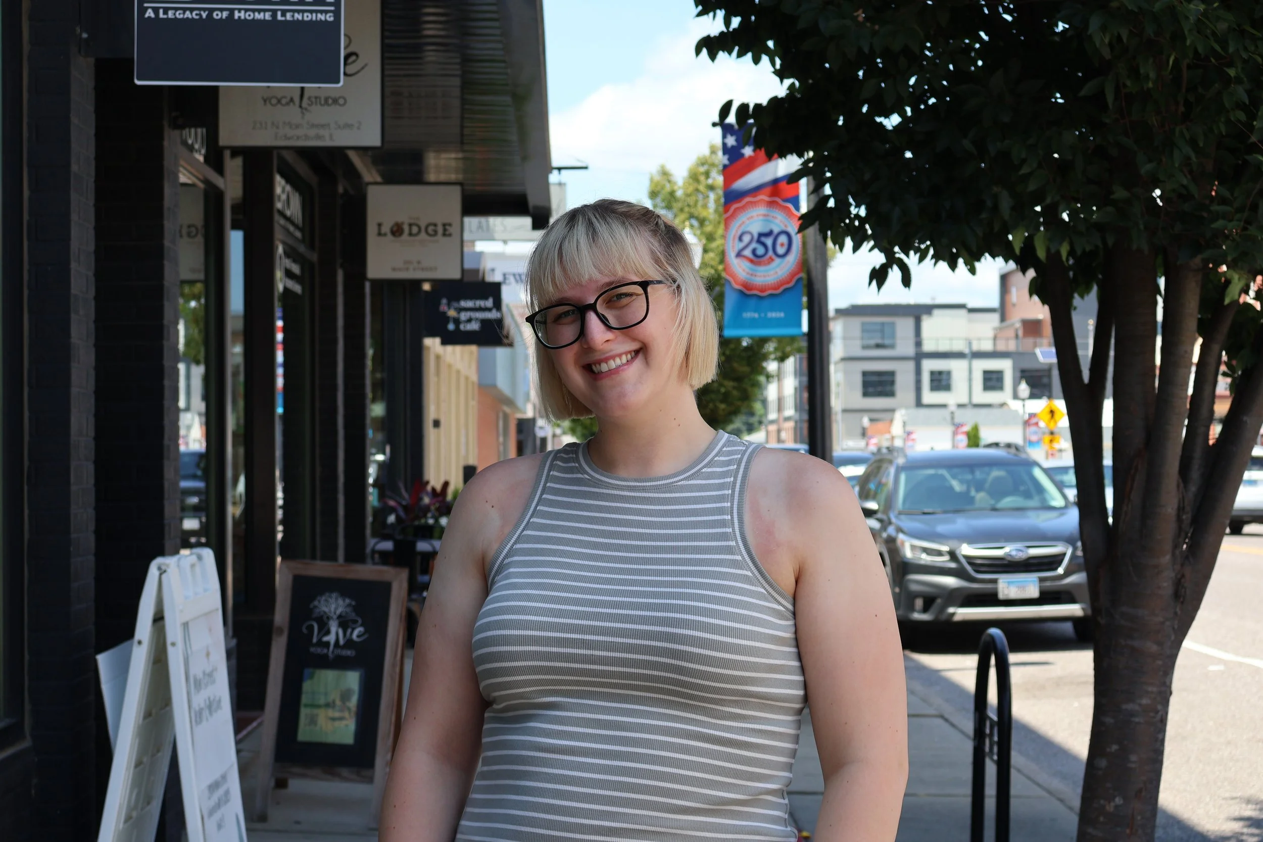 A young woman with blonde hair and black glasses smiling outdoors on a city sidewalk, wearing a sleeveless striped top nearby shops, a tree, and parked cars are visible in the background.