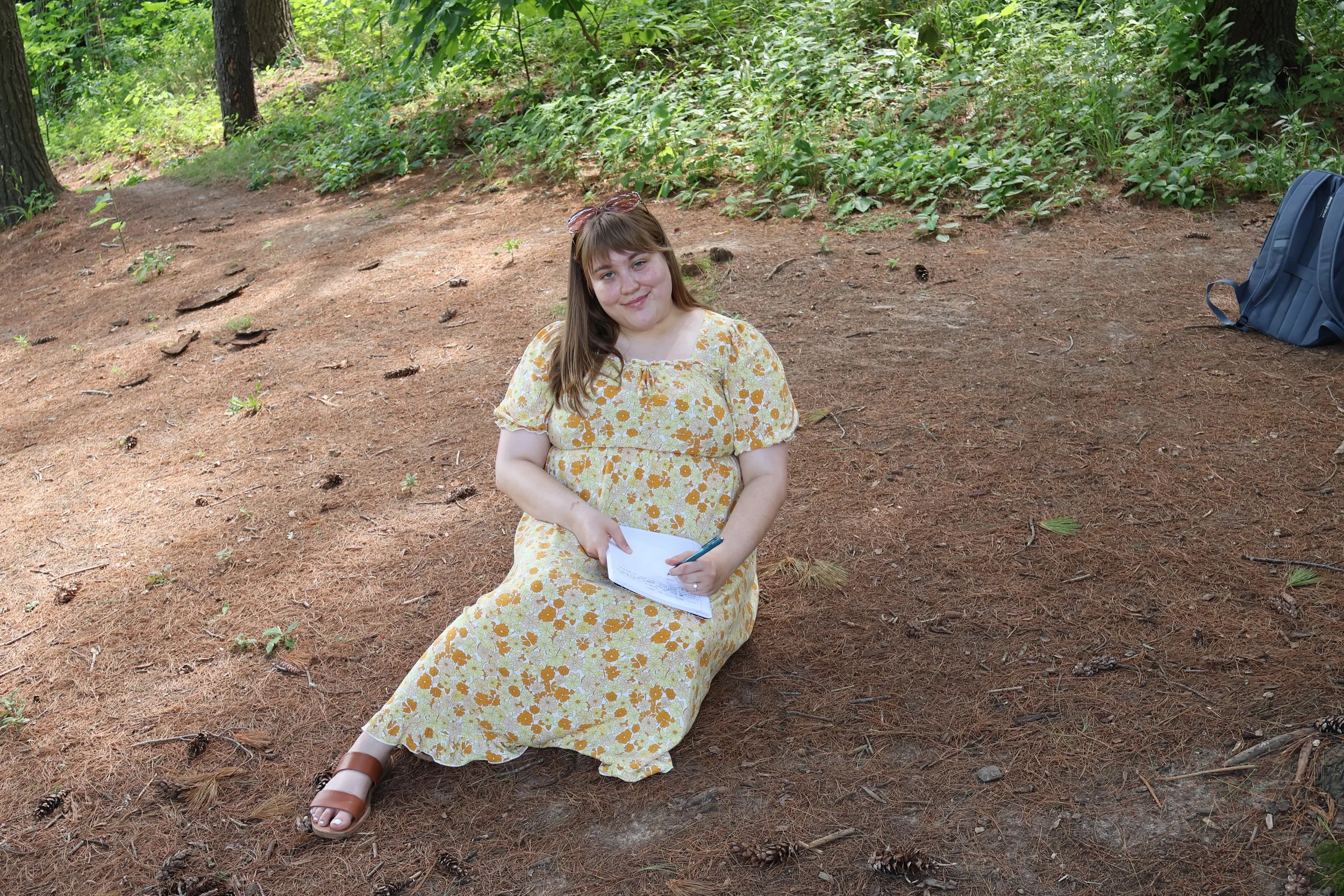 A young woman in a yellow floral dress sitting on the forest floor, holding a notebook and a pen, with trees and green foliage in the background, and a blue backpack to her right.